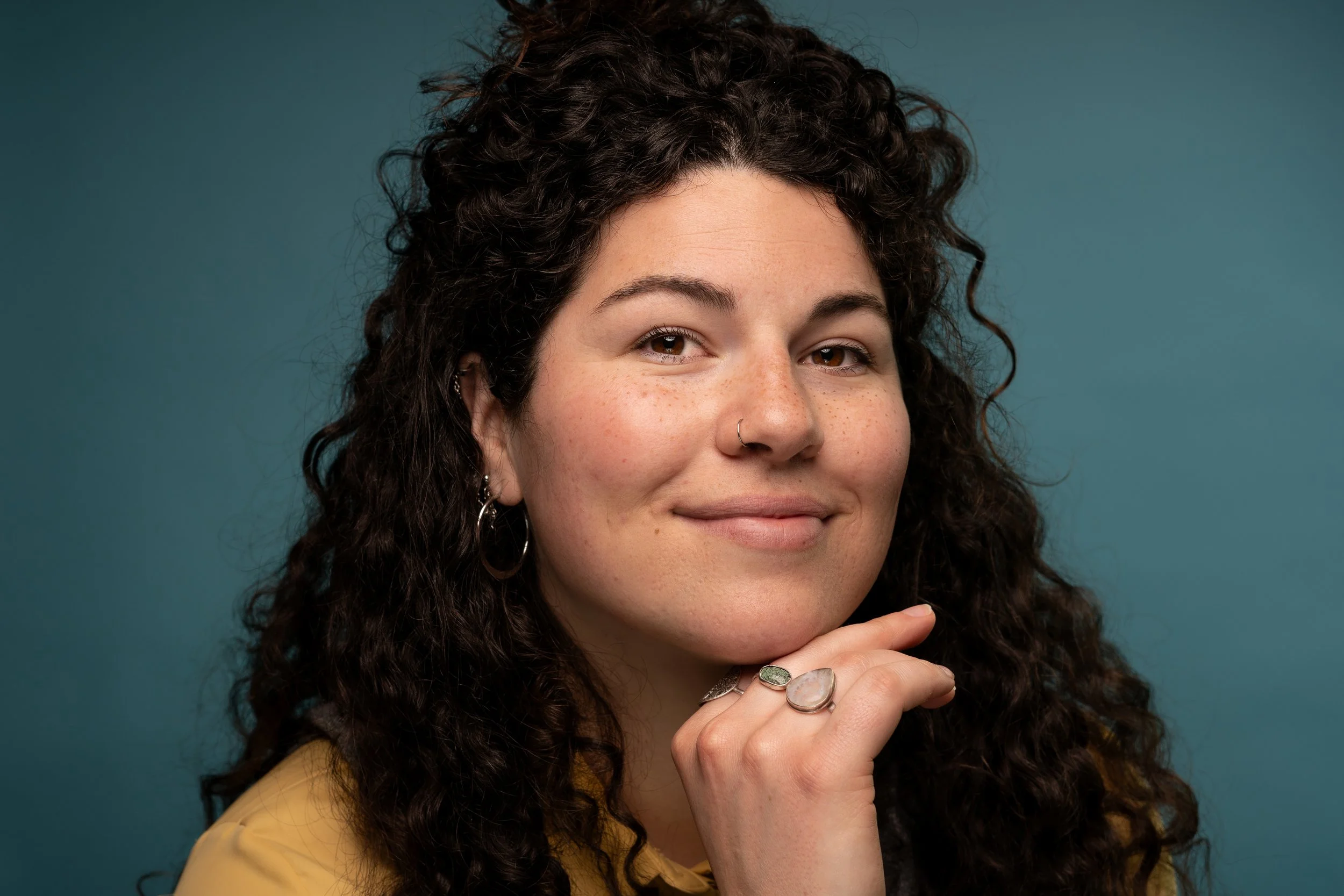 Close-up of a woman with curly dark hair, wearing rings and earrings, smiling gently against a teal background.