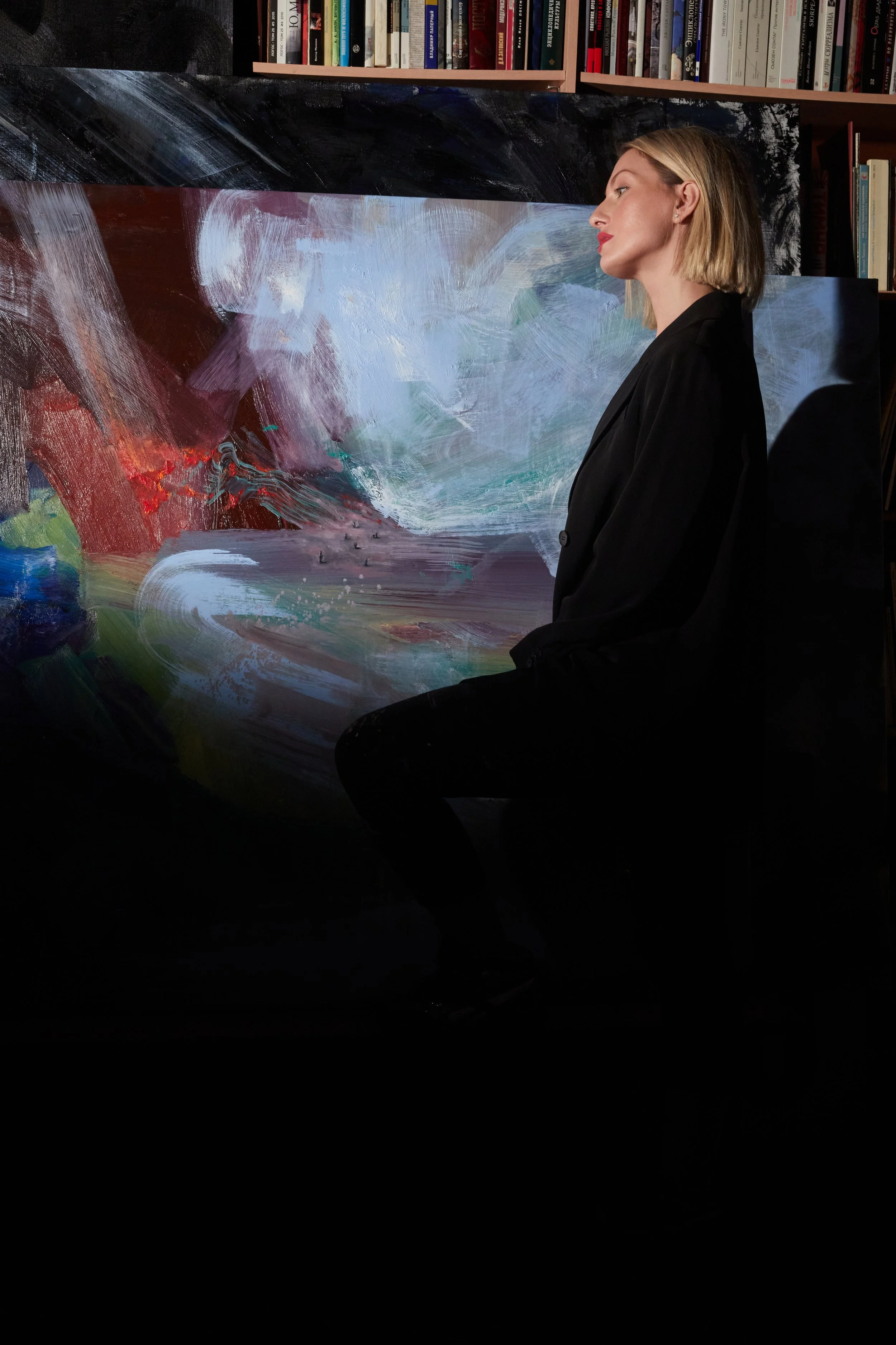 Blonde woman in black standing beside a large abstract painting in a room lined with bookshelves