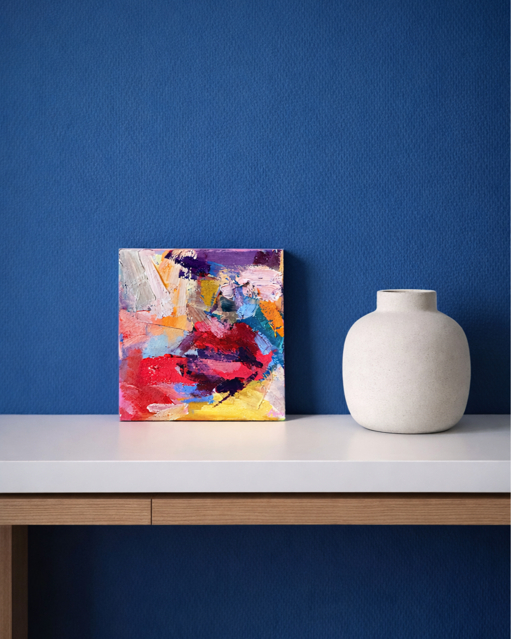 Colorful abstract painting and white ceramic vase on a modern console table against a blue wall