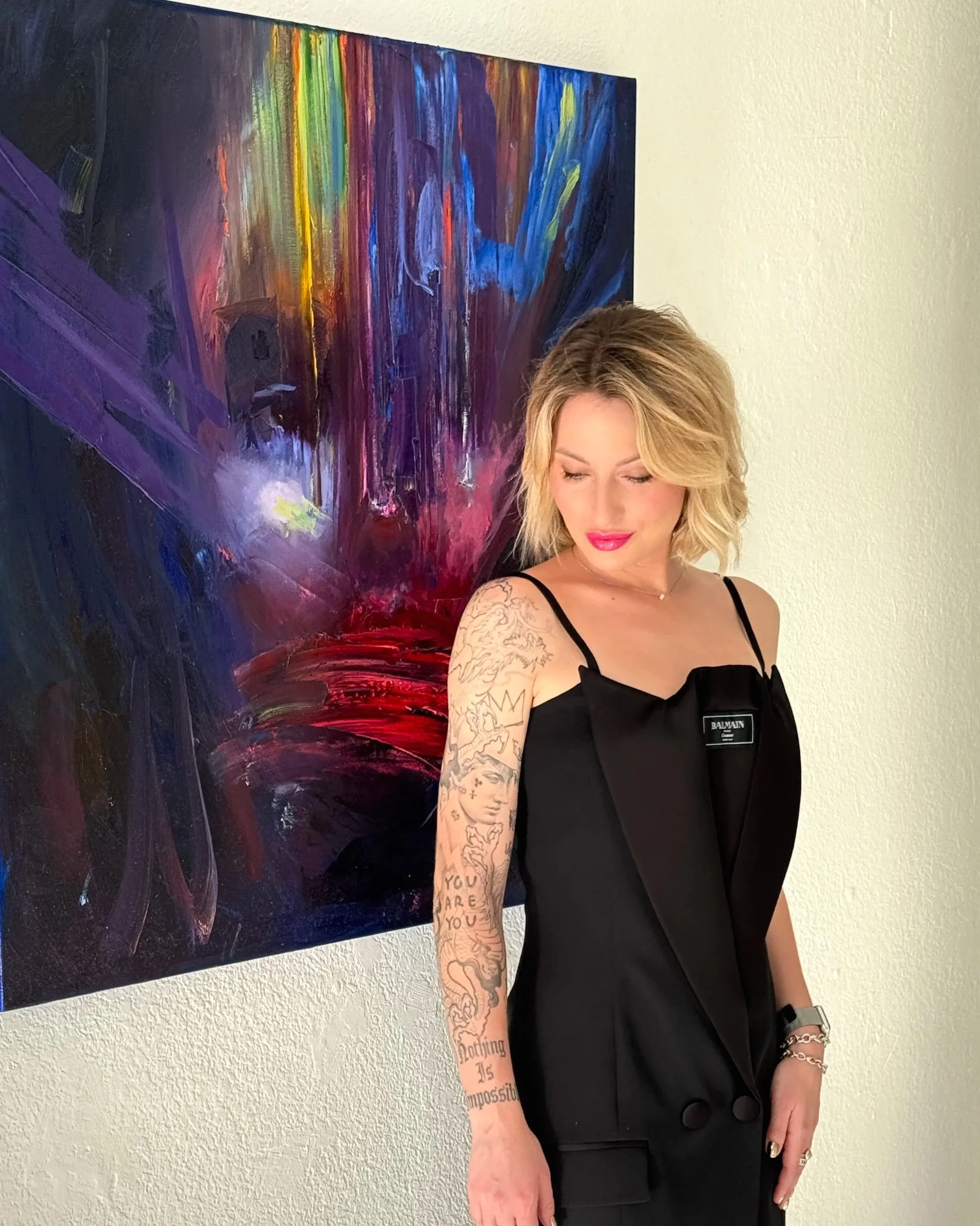 Blonde tattooed woman in a black Balmain outfit standing beside a colorful abstract painting.