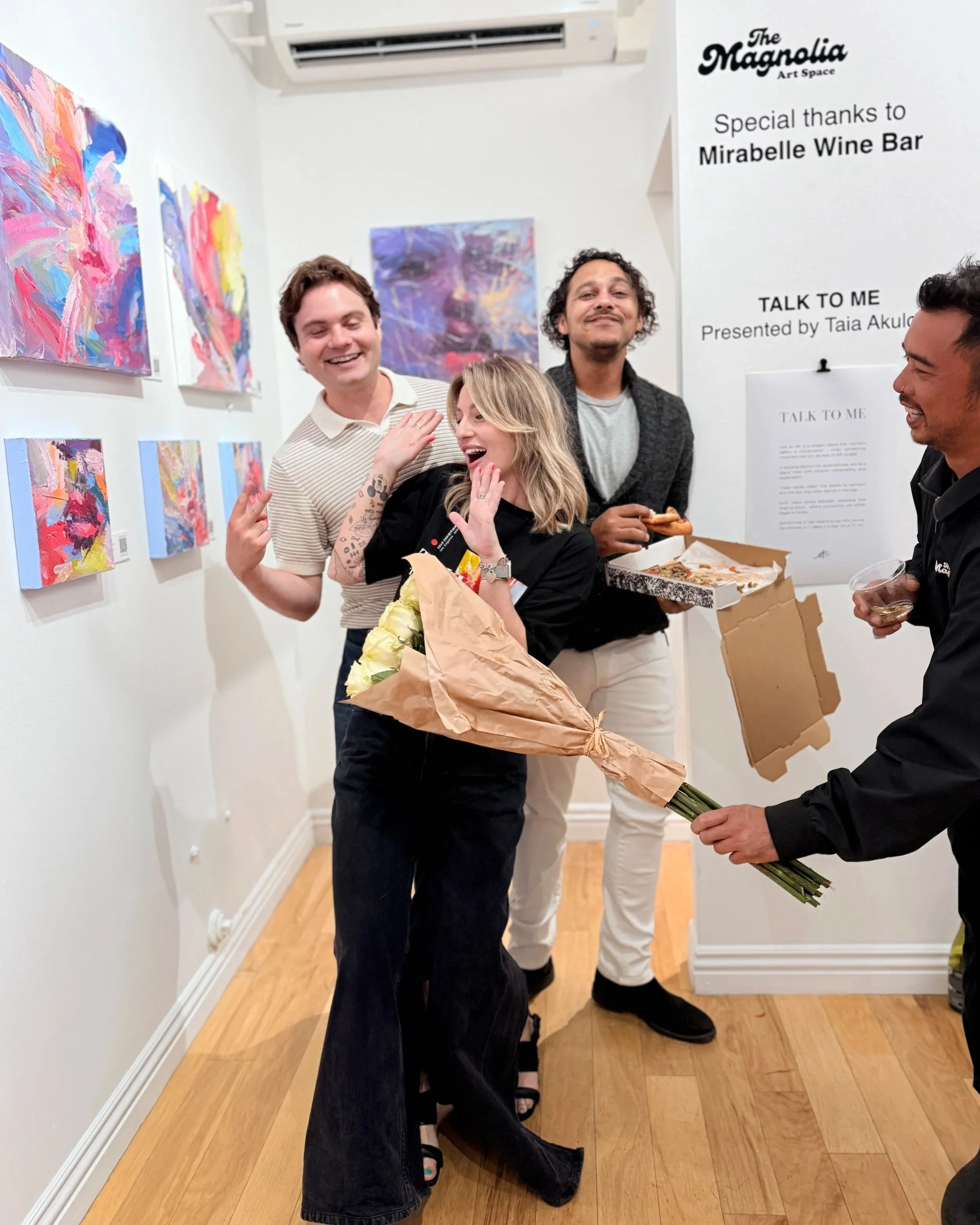 Group of people celebrating in an art gallery surrounded by colorful abstract paintings, flowers, food, and wine.