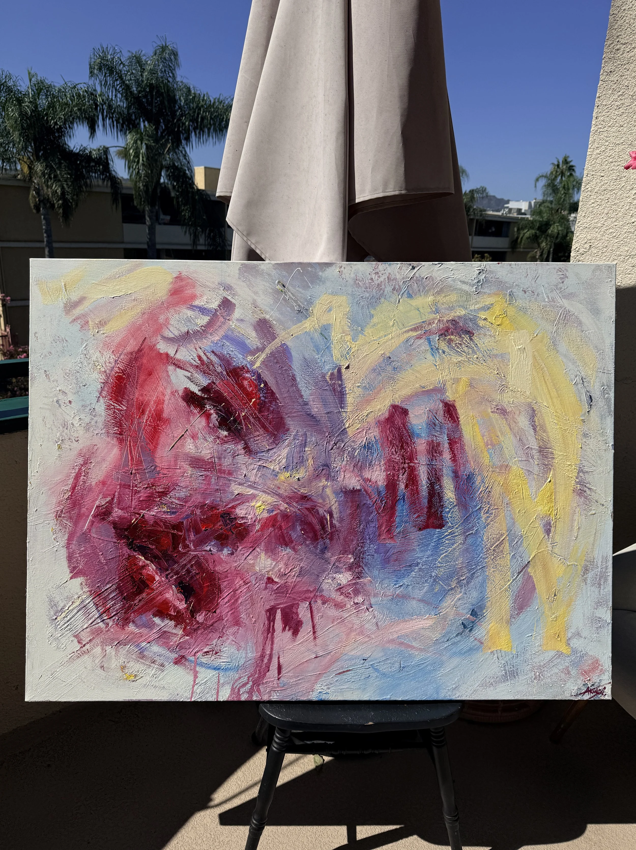 Colorful abstract textured painting on an easel outdoors, with a patio umbrella, palm trees, and blue sky in the background.