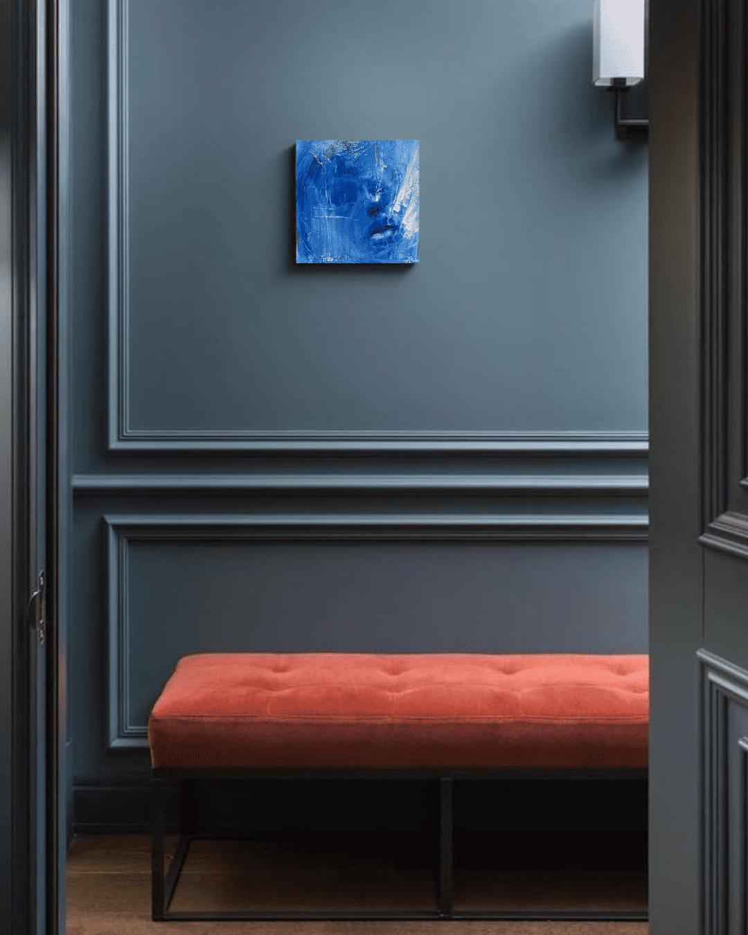 Red upholstered bench beneath a blue abstract painting in a dark blue paneled room