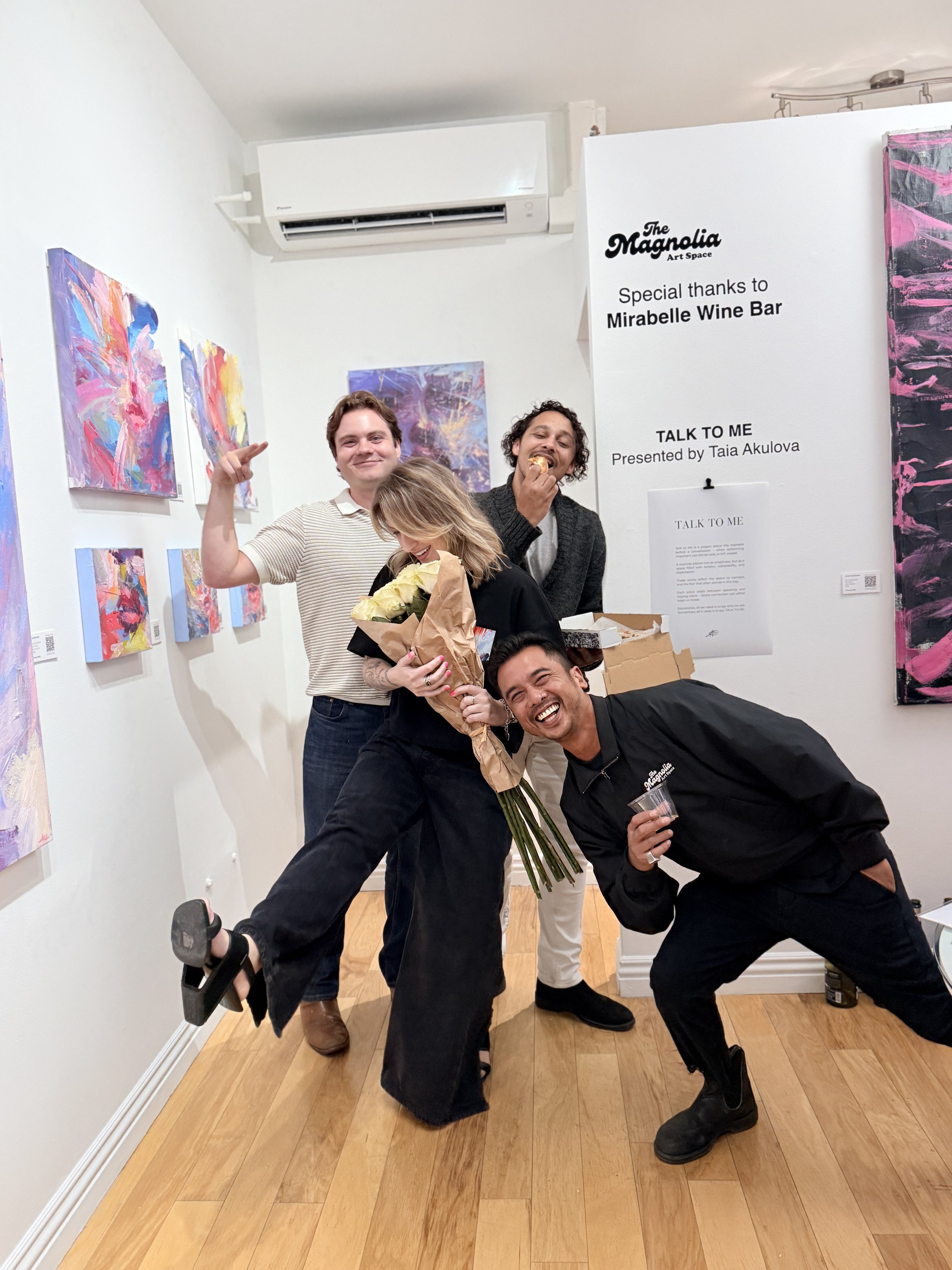 Four people pose playfully at The Magnolia Art Space beside colorful abstract paintings and an exhibition sign.