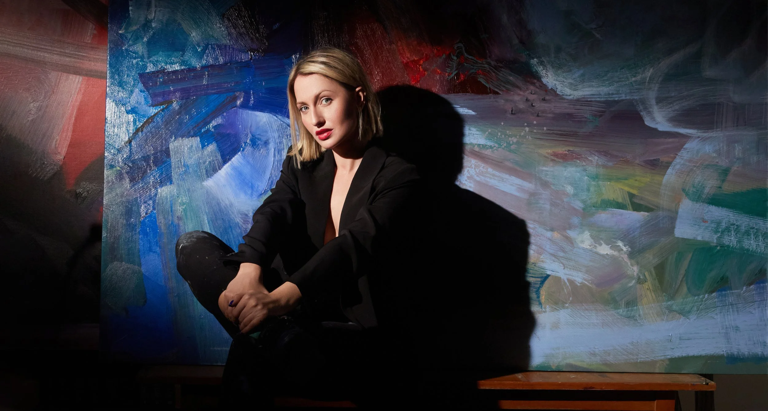 Blonde woman in a black outfit seated in front of a colorful abstract painting