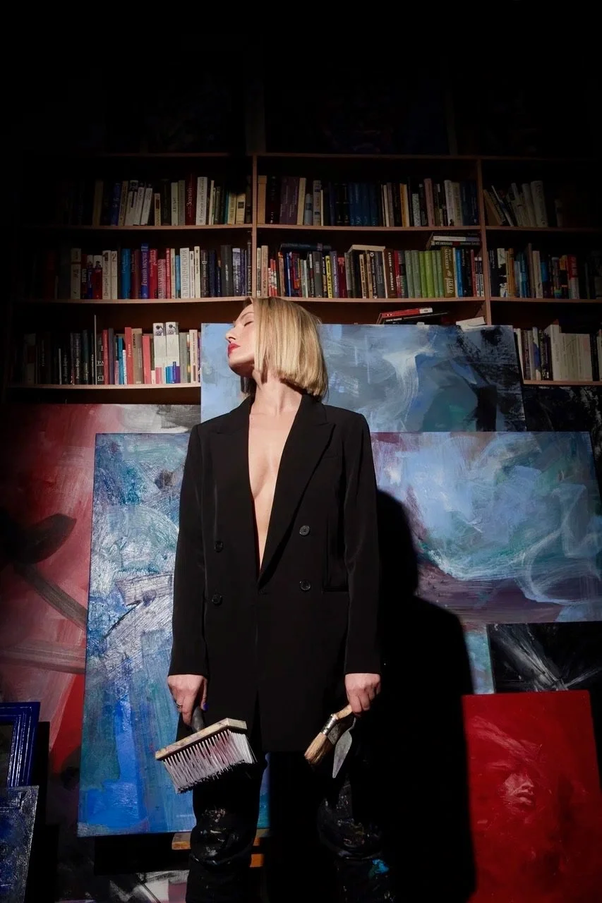Blonde woman in a black suit holding paintbrushes in front of abstract paintings and a bookshelf