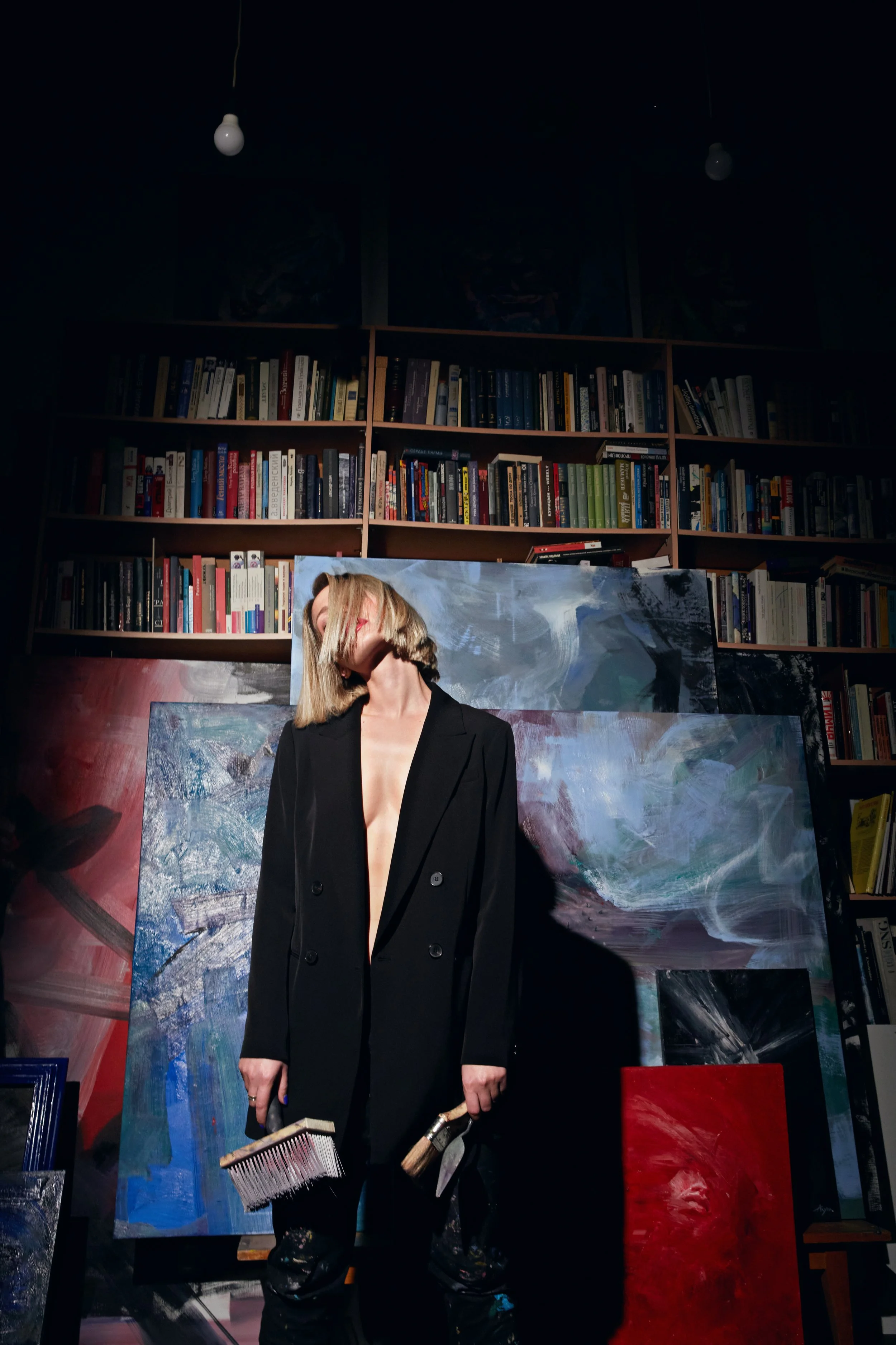 Artist in a black suit holding paintbrushes in front of abstract paintings and a bookshelf