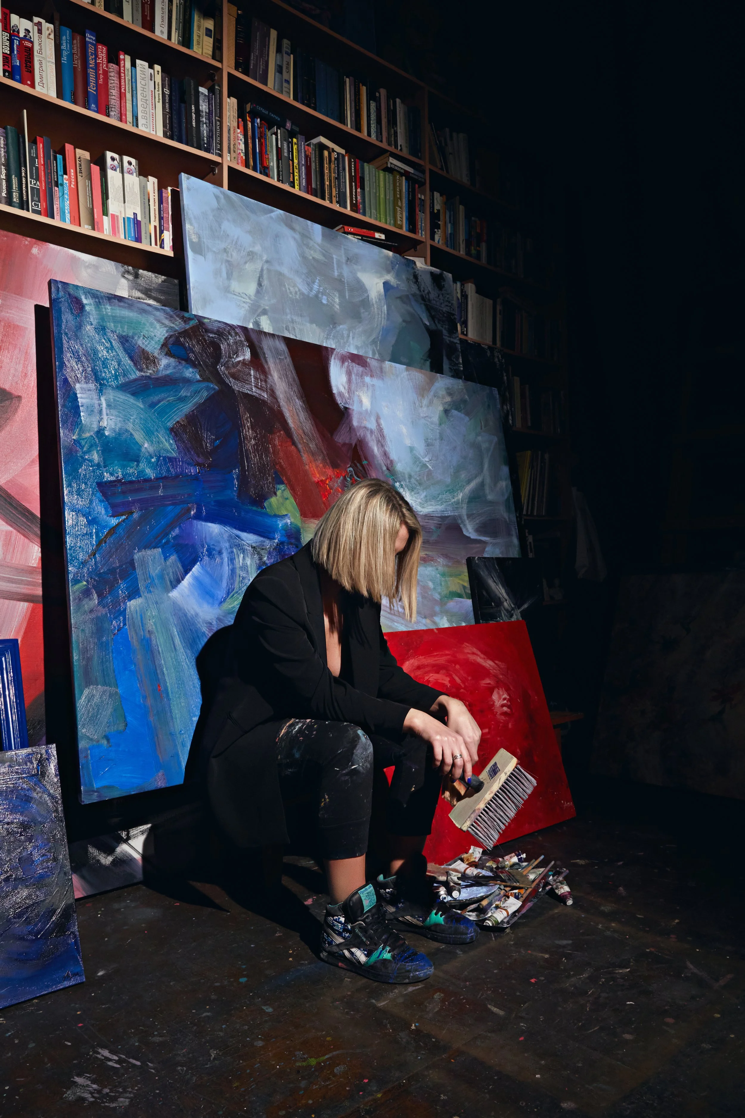 Artist in black clothing holding a paintbrush beside colorful abstract paintings and bookshelves in a dark studio