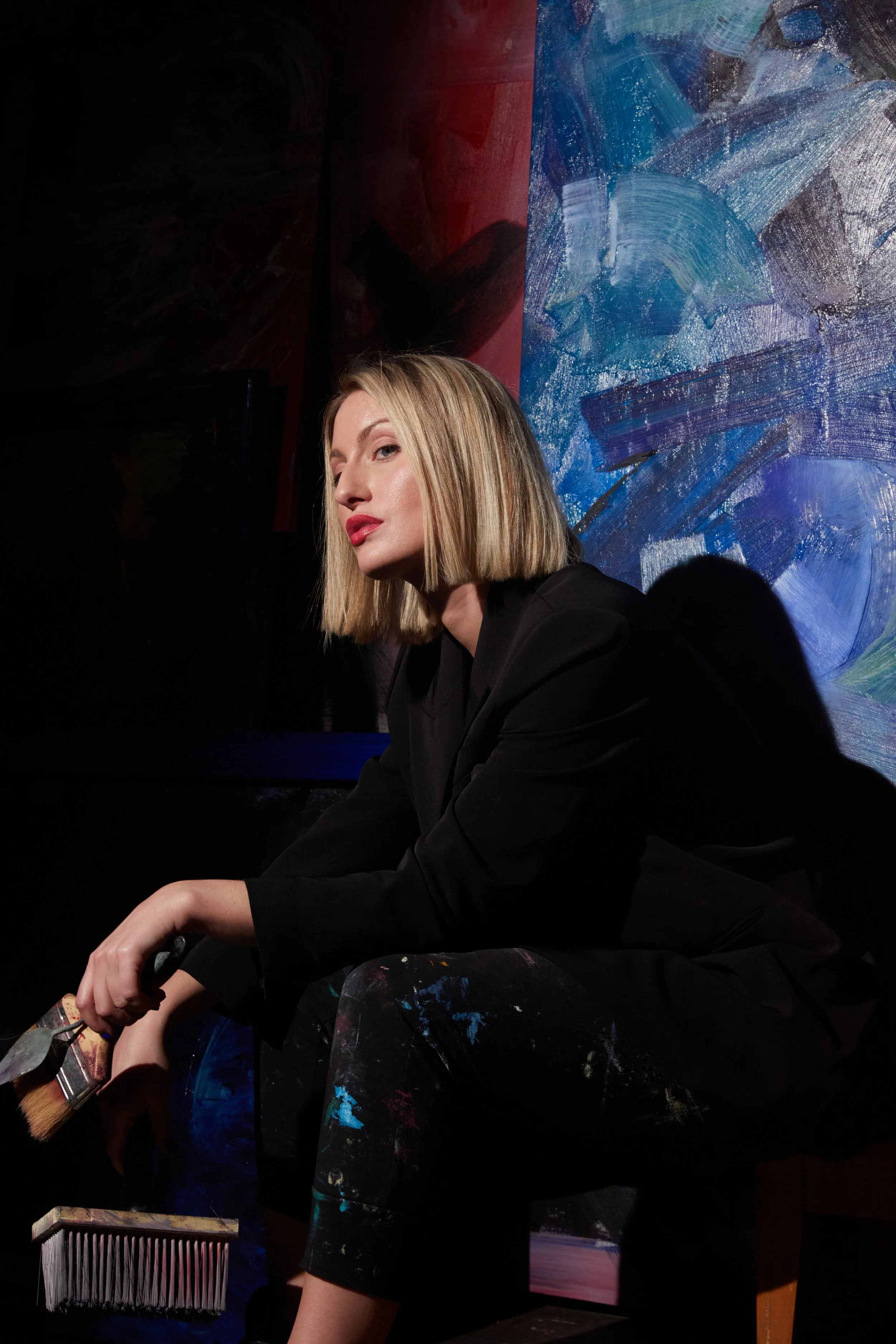 Blonde woman in black clothing holding a paintbrush in front of a colorful abstract painting.