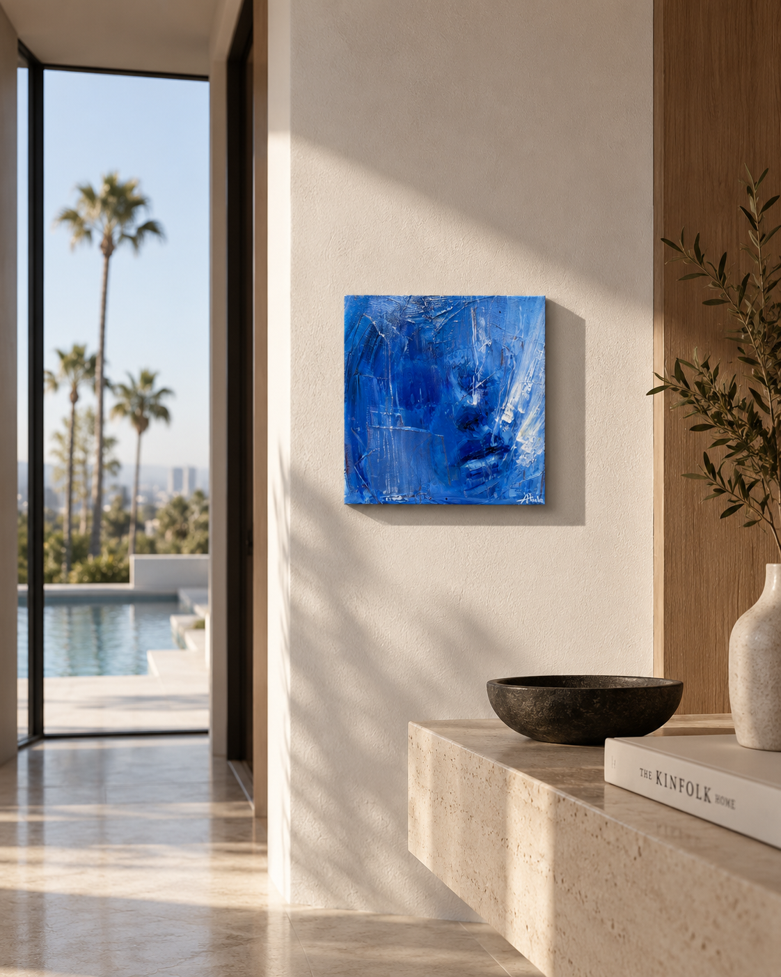 Modern sunlit interior with blue abstract wall art, travertine console, black bowl, olive branch vase, and poolside palm view