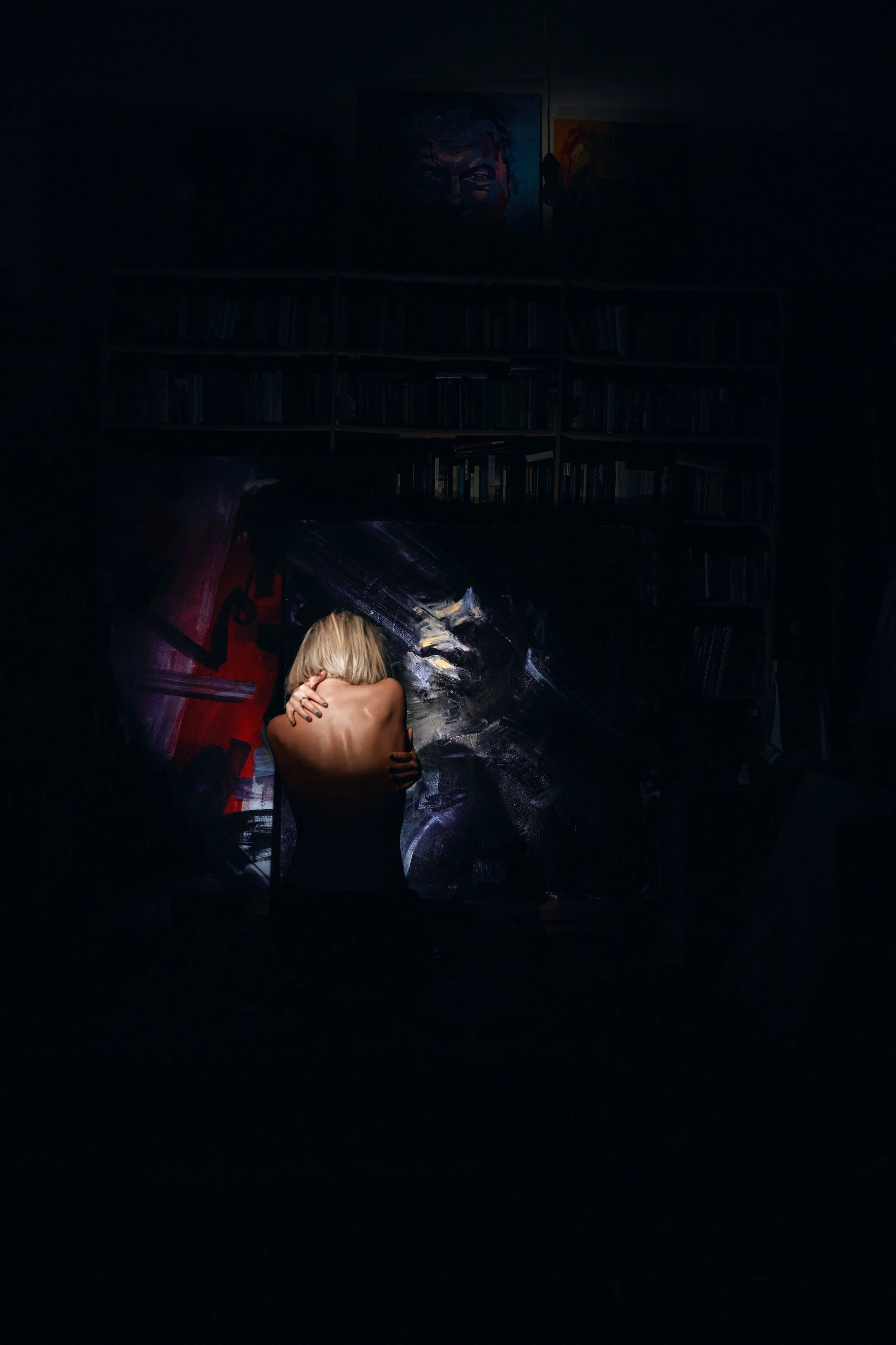 Dark artistic scene of a blonde woman embracing a large black-and-white painted figure in front of bookshelves and portraits