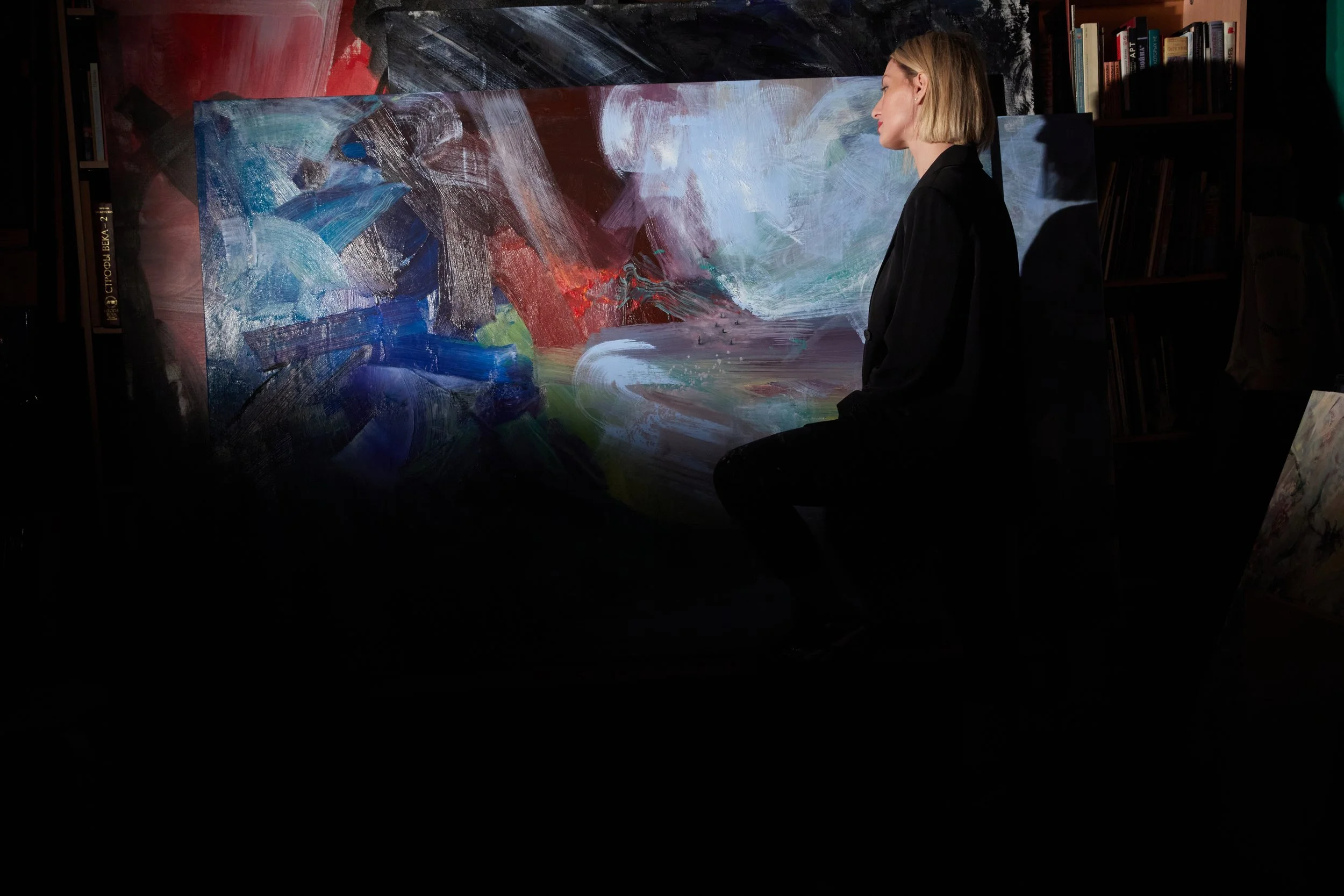 Woman in black clothing standing beside a large abstract painting in a dimly lit art studio with bookshelves.