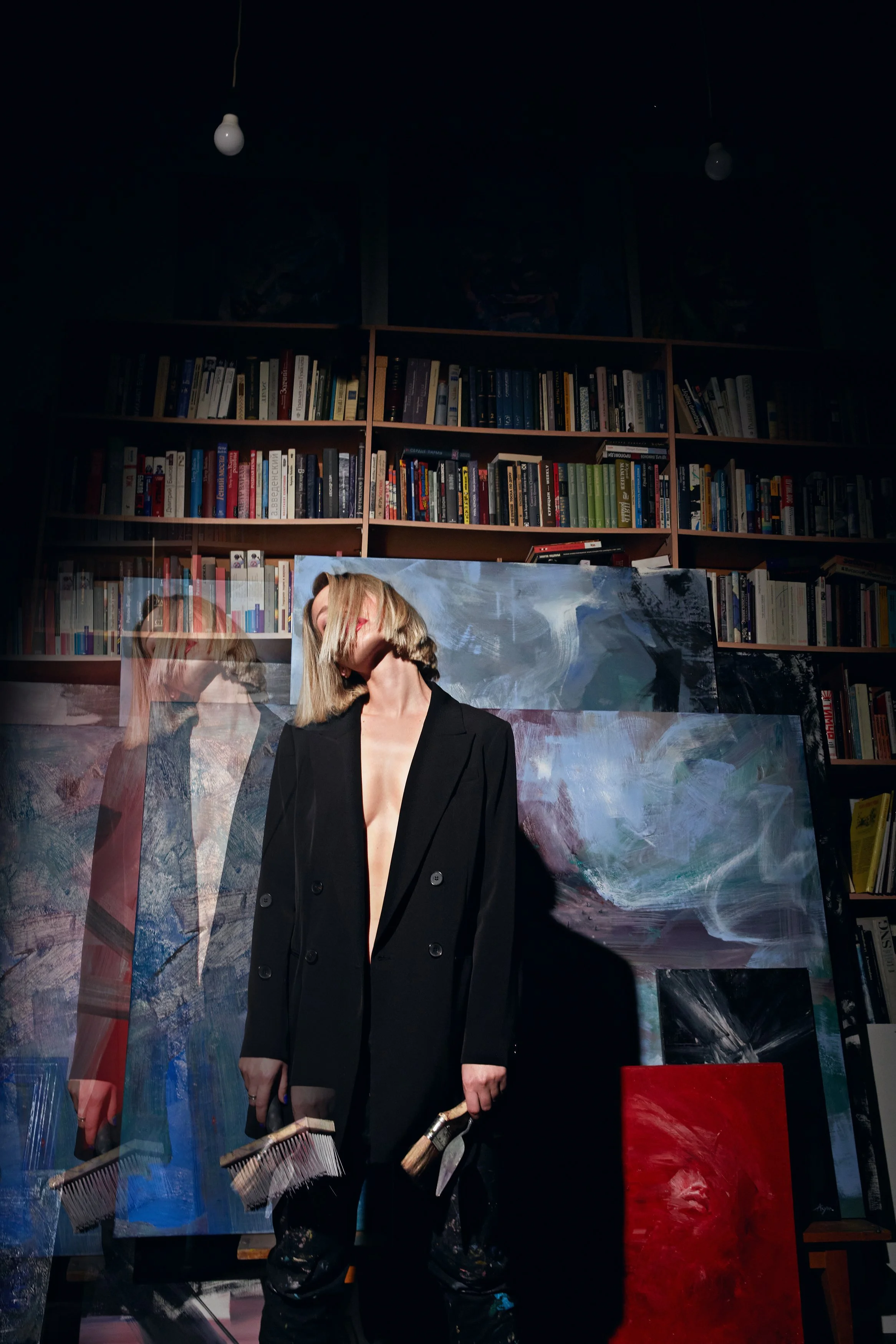 Artist in black clothing holding paintbrushes in front of abstract paintings and bookshelves