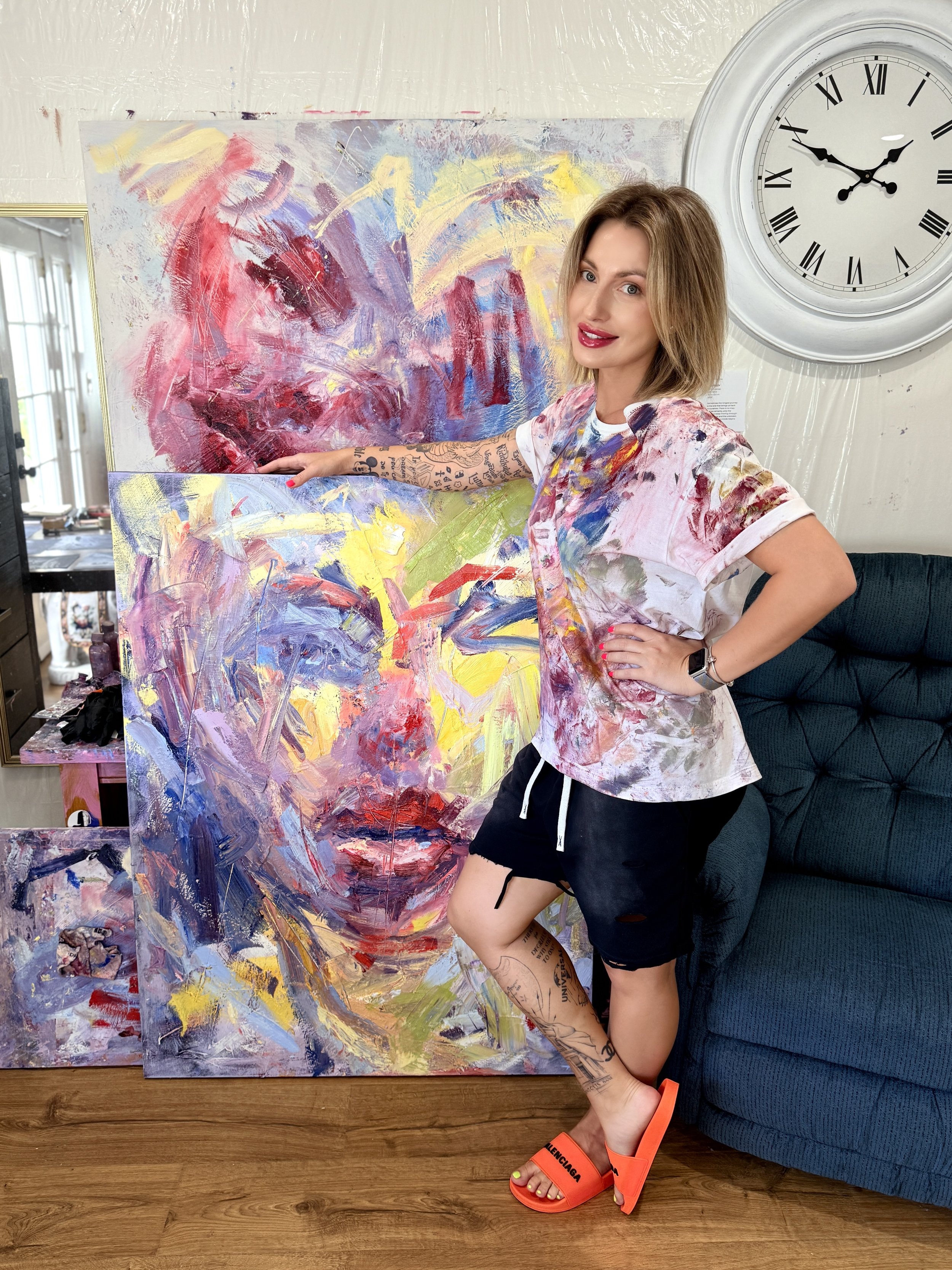 Woman posing beside a large colorful abstract painting in a home studio, with a wall clock and additional artwork in the background.