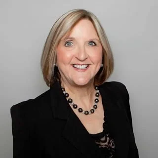 Carol Brenstuhl smiling with shoulder-length blonde hair, wearing a black blazer, black necklace, and a black top, against a light gray background.