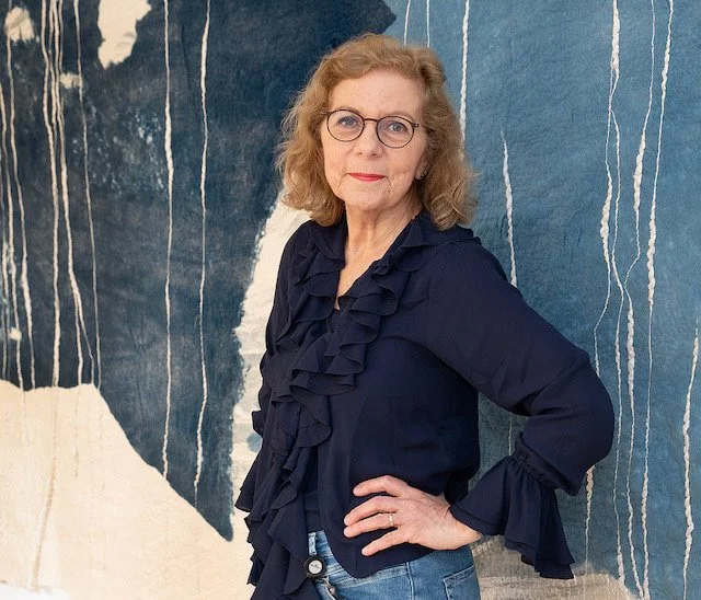 Een vrouw met bril en gekruld, lichtbruin haar poseert tegen een blauwe wand met witte strepen, gekleed in een donkerblauwe blouse met ruffles en blauwe jeans.