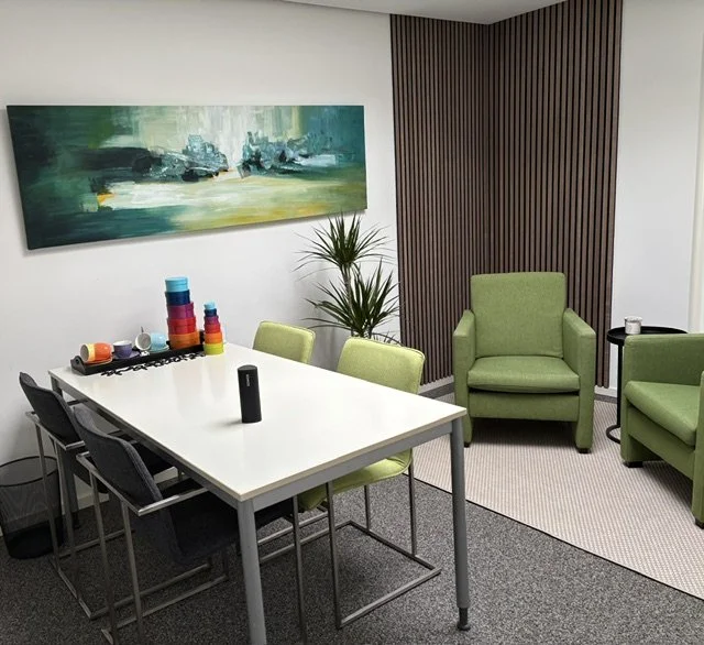 Een vergaderruimte met een witte coachtafel, limekleurige en grijze stoelen, groene fauteuils, een plant, een muur met houten lamellen, en een abstract schilderij.