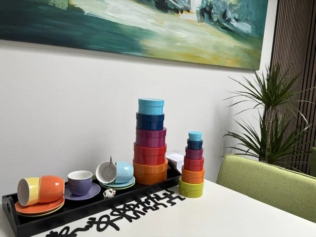 Gekleurde kopjes, schotels en gestapelde doosjes op een tafel, met een plant op de achtergrond.
