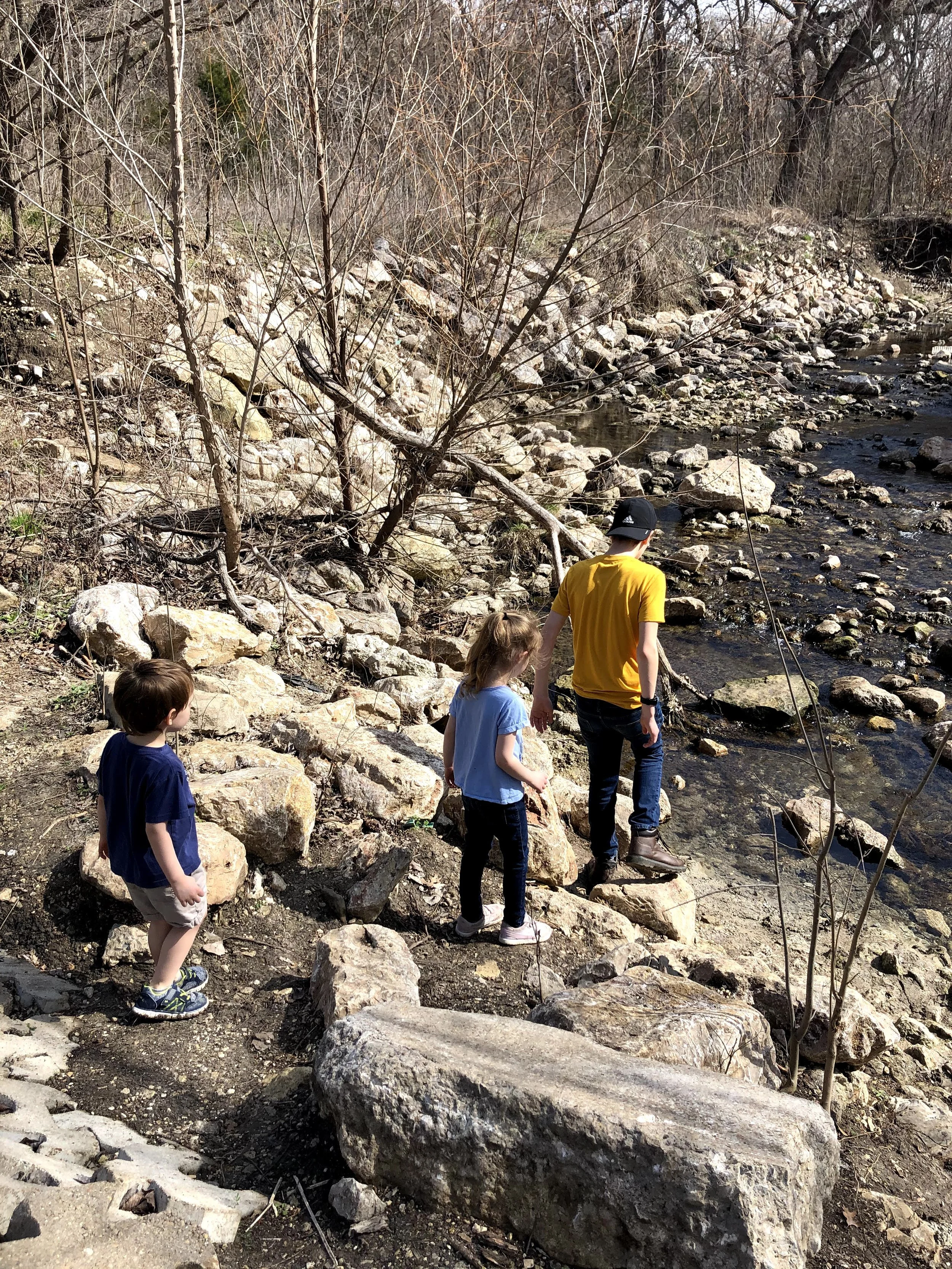 A group of children and a teenager by a rocky riverbank, some looking at the water, some walking along the rocks, with leafless trees and bushes in the background.