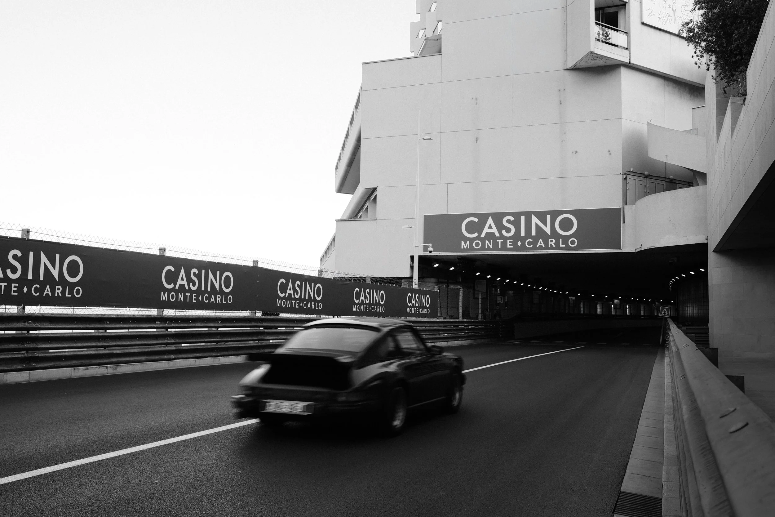 Black-and-white automotive photograph of a classic Porsche 911 entering the Monaco Grand Prix tunnel beneath Casino Monte-Carlo signage.