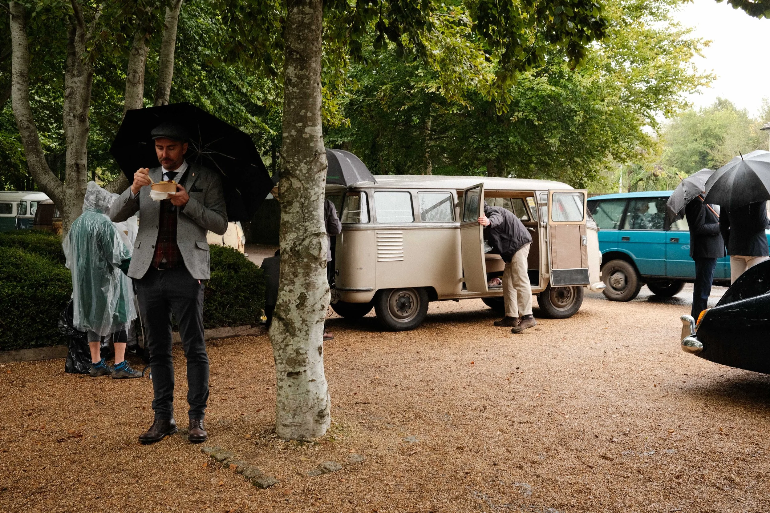 Vintage Volkswagen camper van at the Goodwood Revival in the rain, with visitors in period clothing sheltering beneath umbrellas.