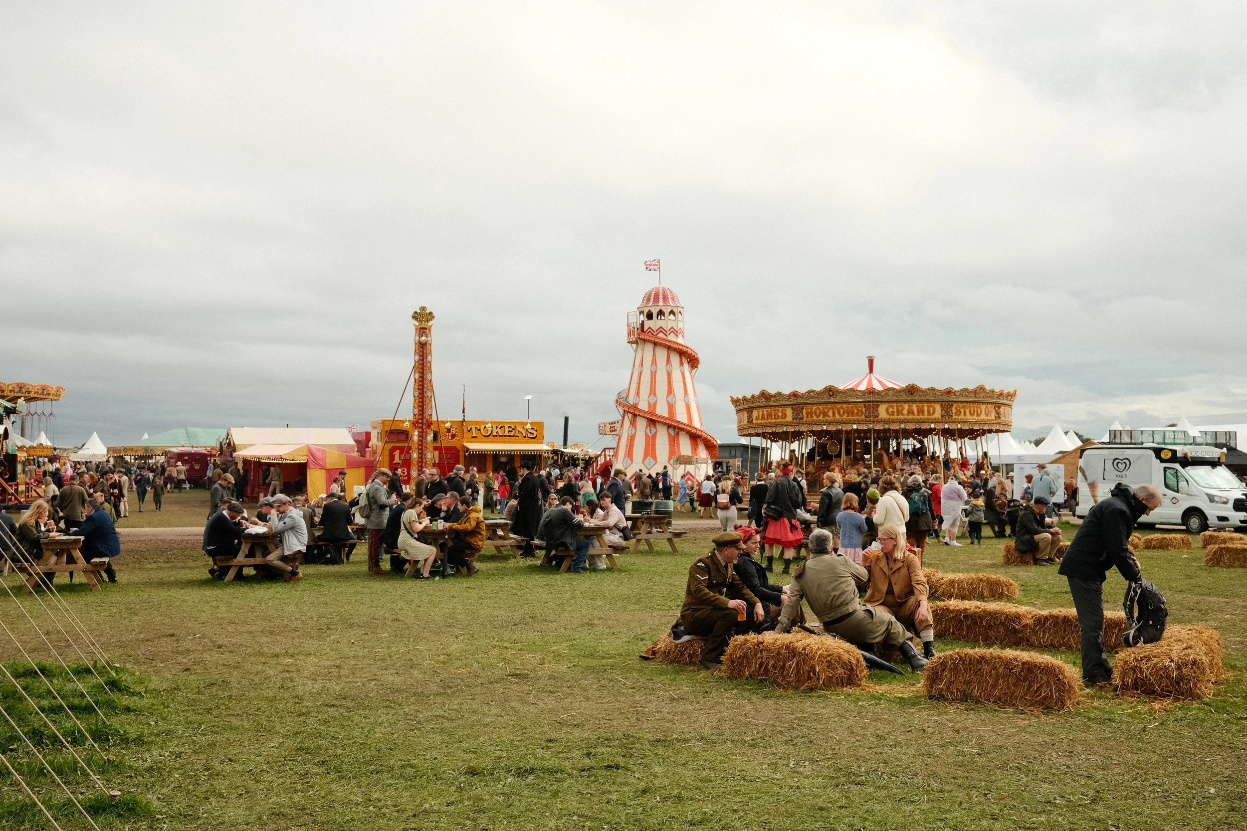 Visitors in period clothing around the vintage fairground at the Goodwood Revival, with carousel rides and hay bale seating beneath an overcast sky.