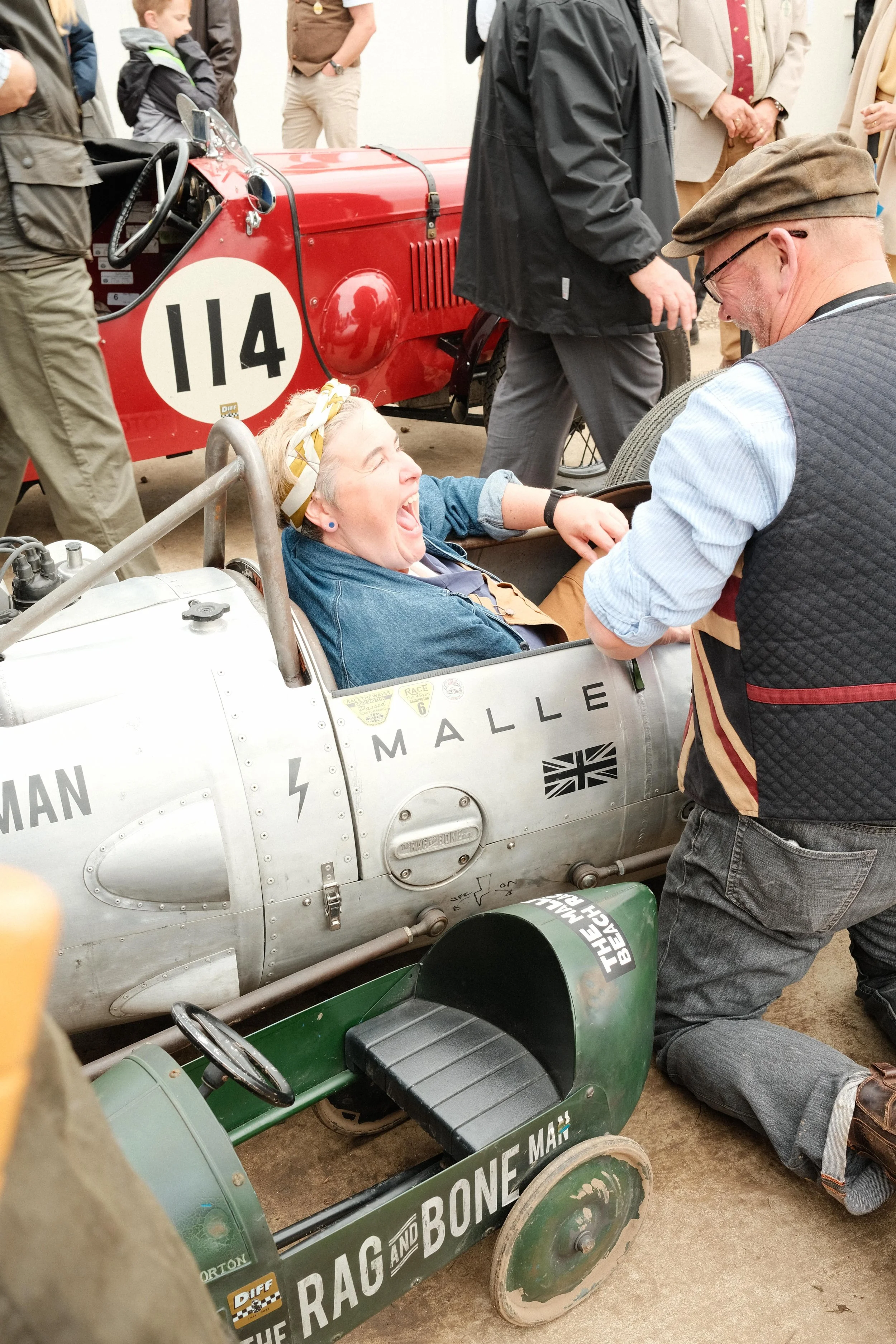 Man and woman laughing beside a small vintage-style race car at the Goodwood Revival, surrounded by historic racing machinery and period-dressed visitors.