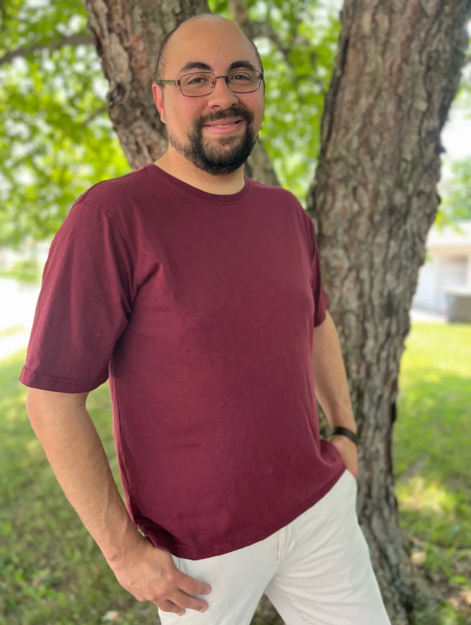 A man with glasses, a beard, and a bald head smiling outdoors next to a tree, wearing a maroon t-shirt and white pants.