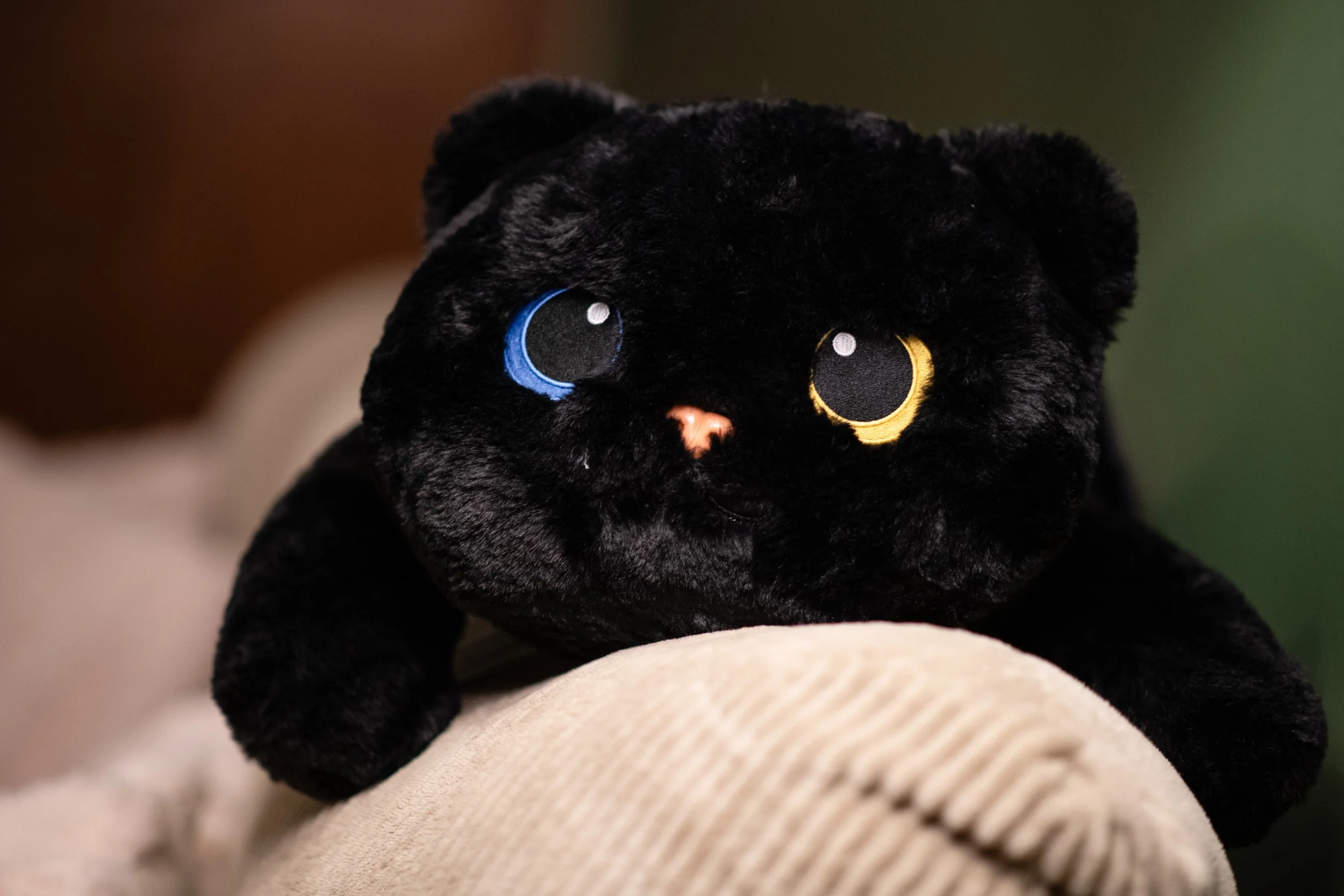 Close-up of a black plush teddy bear with one blue eye and one yellow eye, resting on a beige textured fabric.