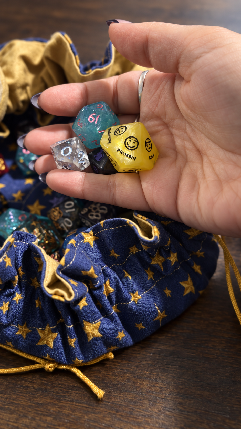Hand holding several colorful polyhedral dice, including a yellow d20 with 'Pleasant' on it, against a star-printed drawstring bag on a wooden surface.