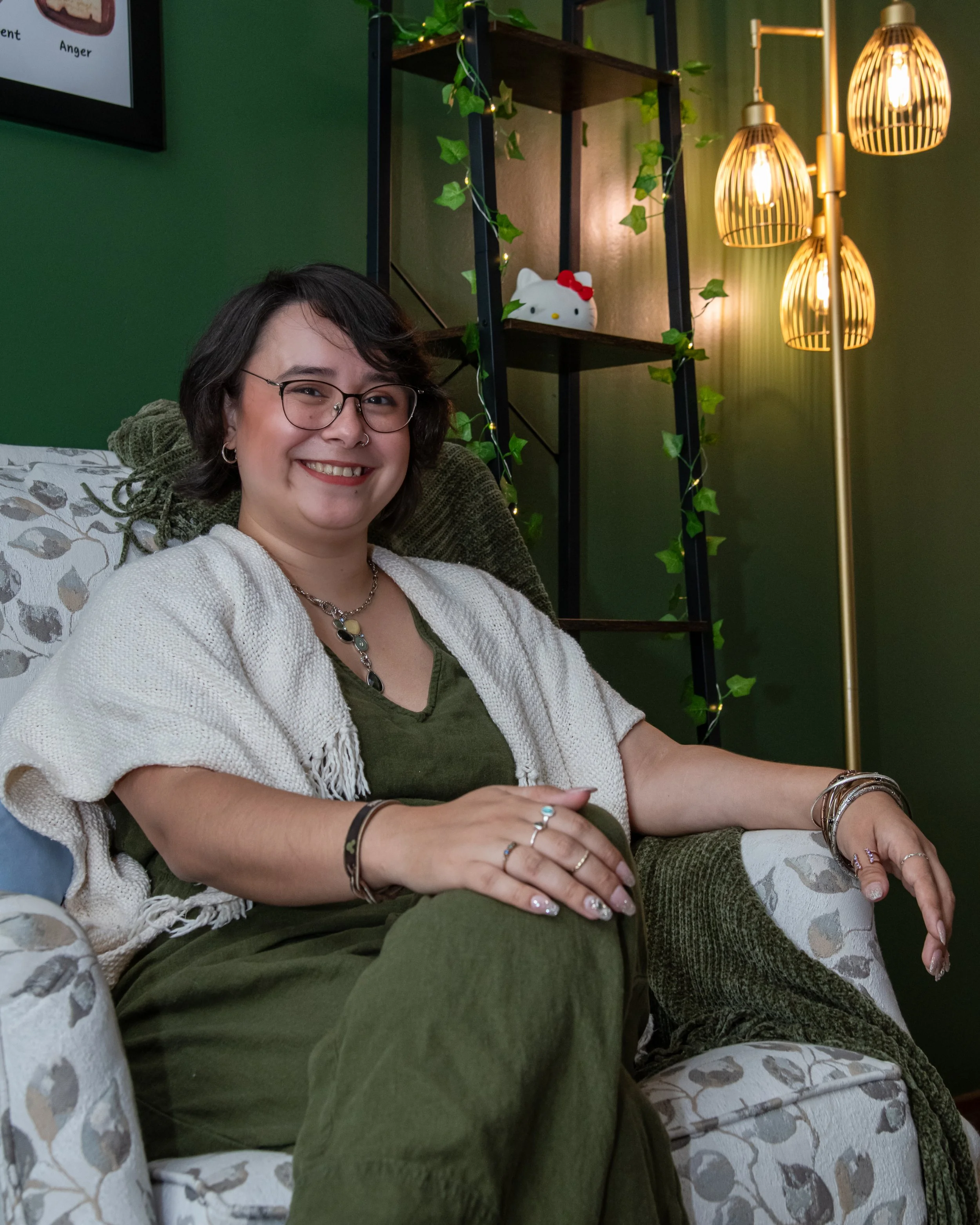 A woman smiling while sitting on a patterned chair in a cozy room with green walls, a bookshelf decorated with green leaves and a Hello Kitty plush toy, and a floor lamp with three warm-colored bulbs.