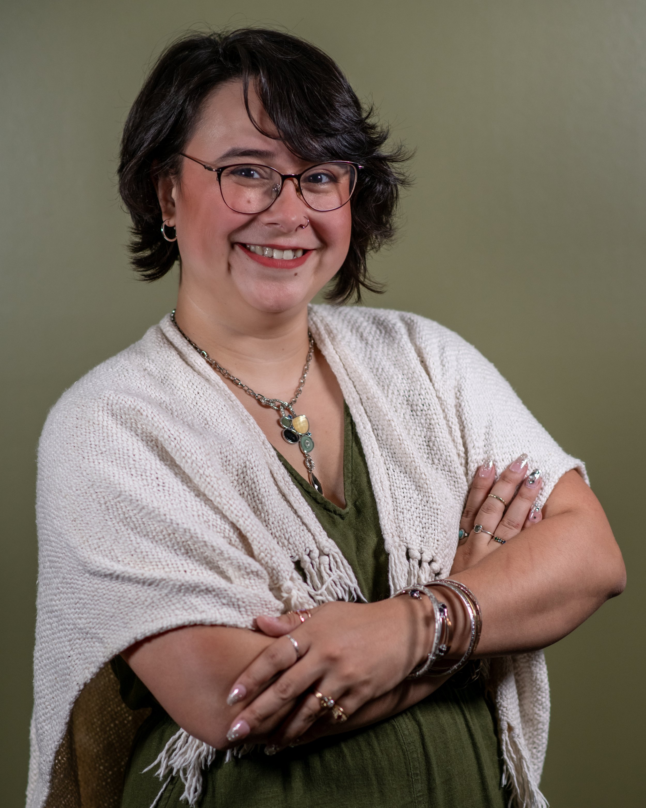 Smiling woman with dark, wavy hair, glasses, and multiple jewelry pieces, crossing her arms in front of a plain, muted background.