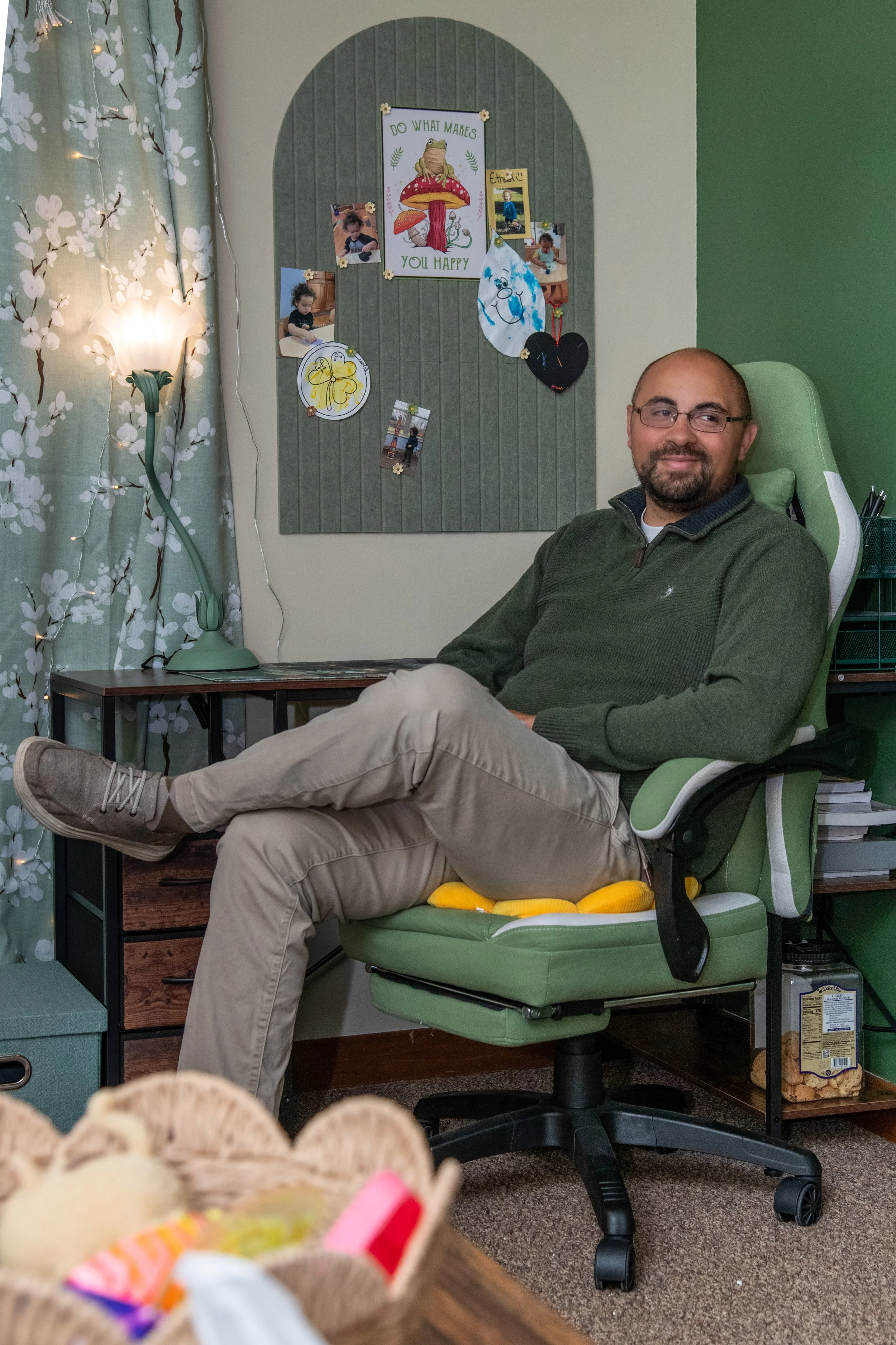 A man with glasses and a beard sitting in a green office chair, smiling, in a room with a decorated bulletin board behind him that has photos and drawings, and a lit lamp on a side table, with a curtain and green and beige walls.
