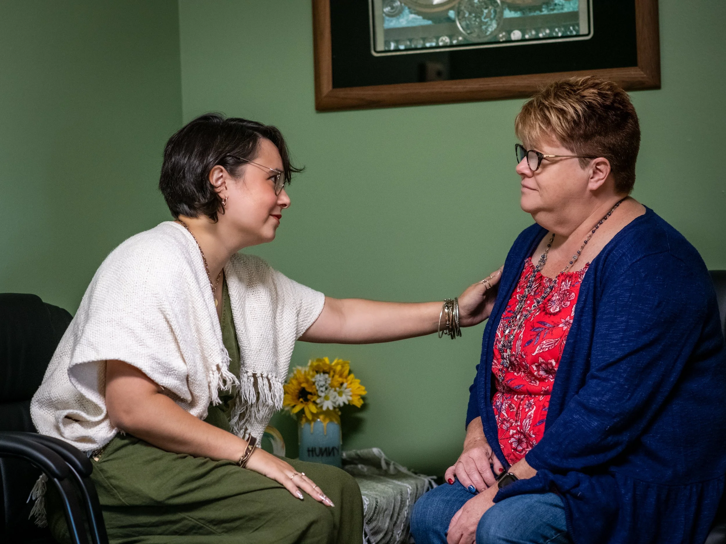 A young woman in a white sweater and green pants is sitting in a wheelchair, facing an older woman with short hair, glasses, and a blue jacket. They are touching hands and sharing a moment of connection in a room with green walls, a flower arrangement, and a framed mirror.