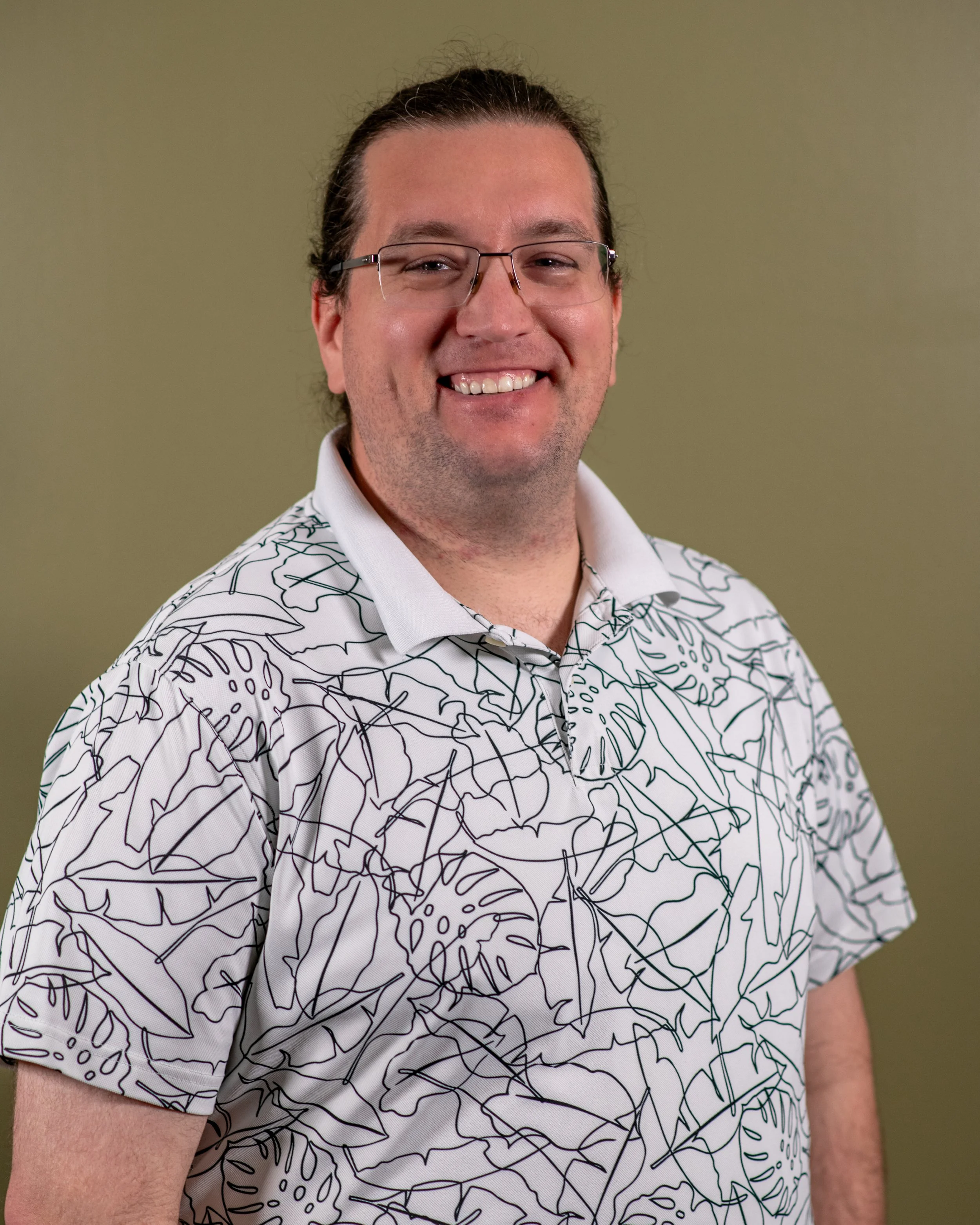 A smiling man with glasses, wearing a white shirt with a black abstract line pattern, standing against a plain greenish background.