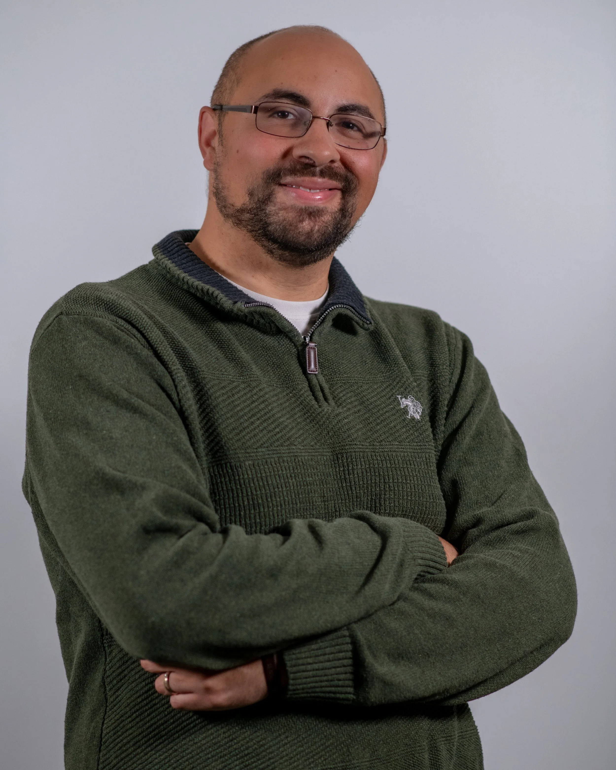 A man with glasses and a goatee, wearing a green zip-up sweater, standing against a plain light gray background.