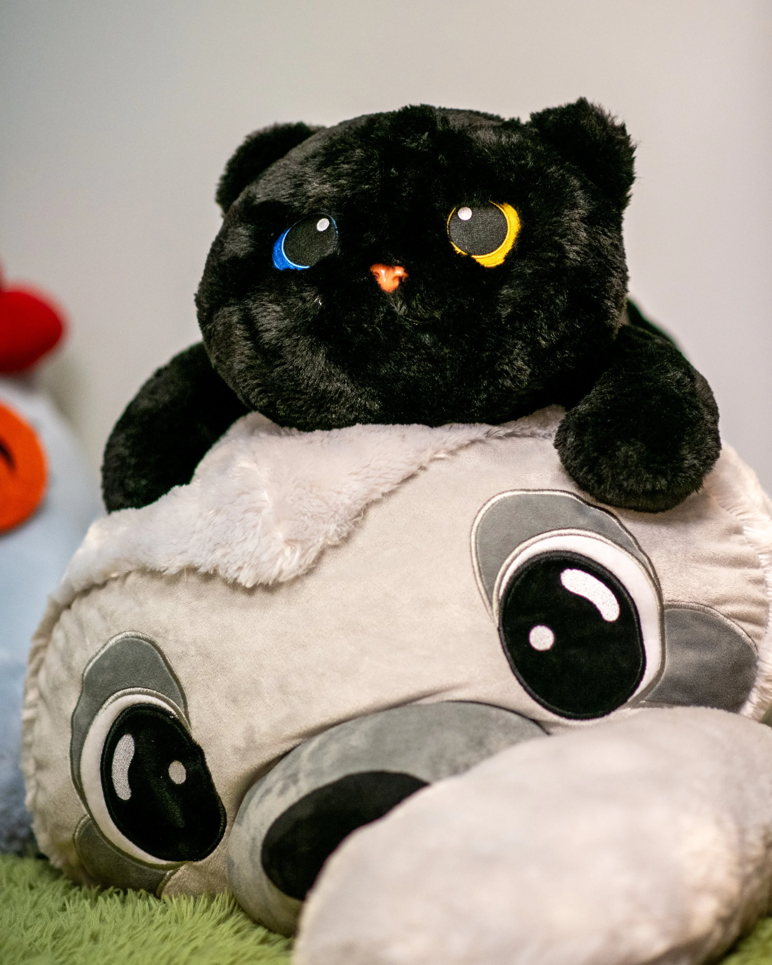 A close-up of a black plush teddy bear with yellow and blue eyes, resting on other plush toys, with a gray background and a green fuzzy surface.