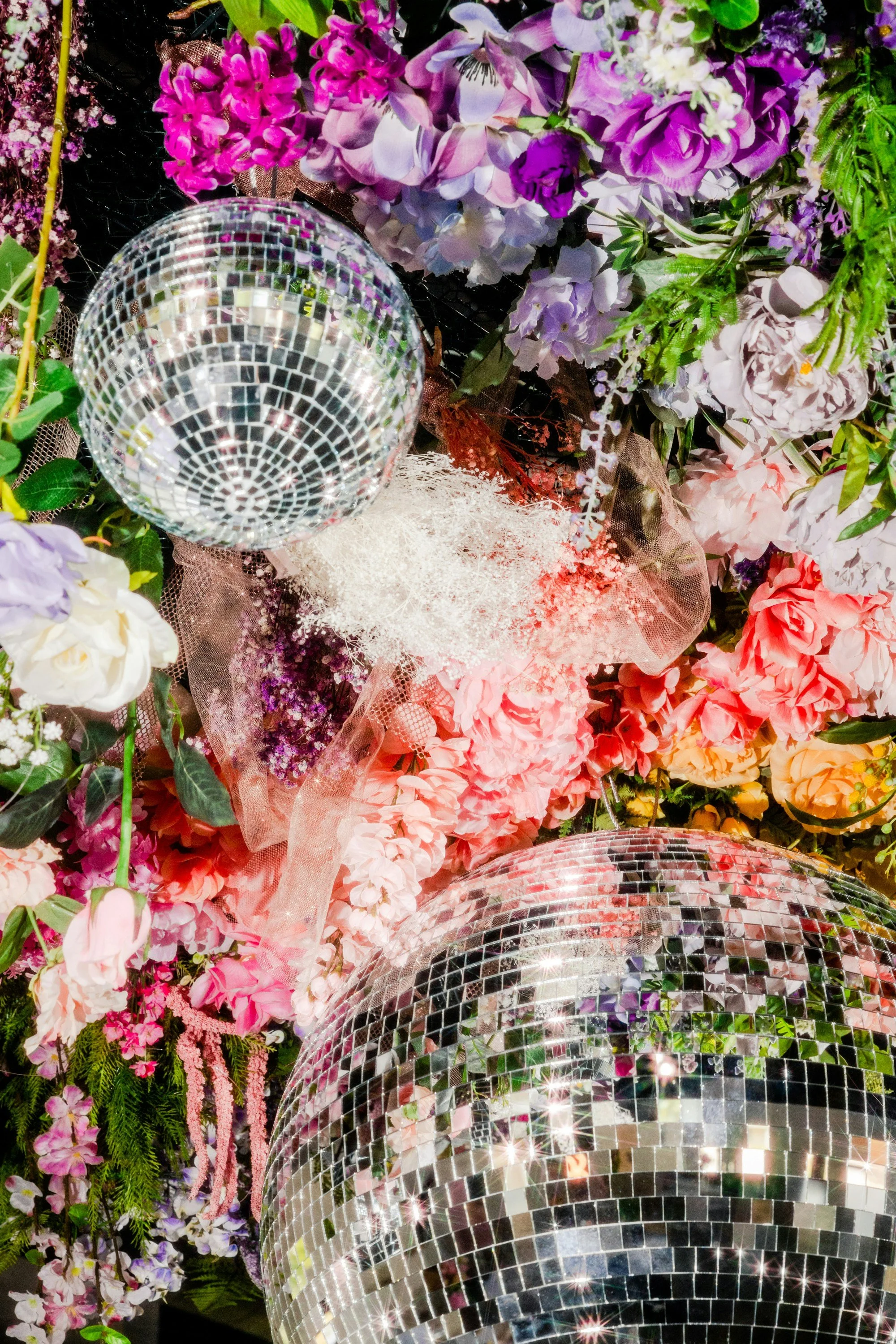 Disco balls hanging amidst a vibrant arrangement of pink, purple, and white flowers with green foliage.