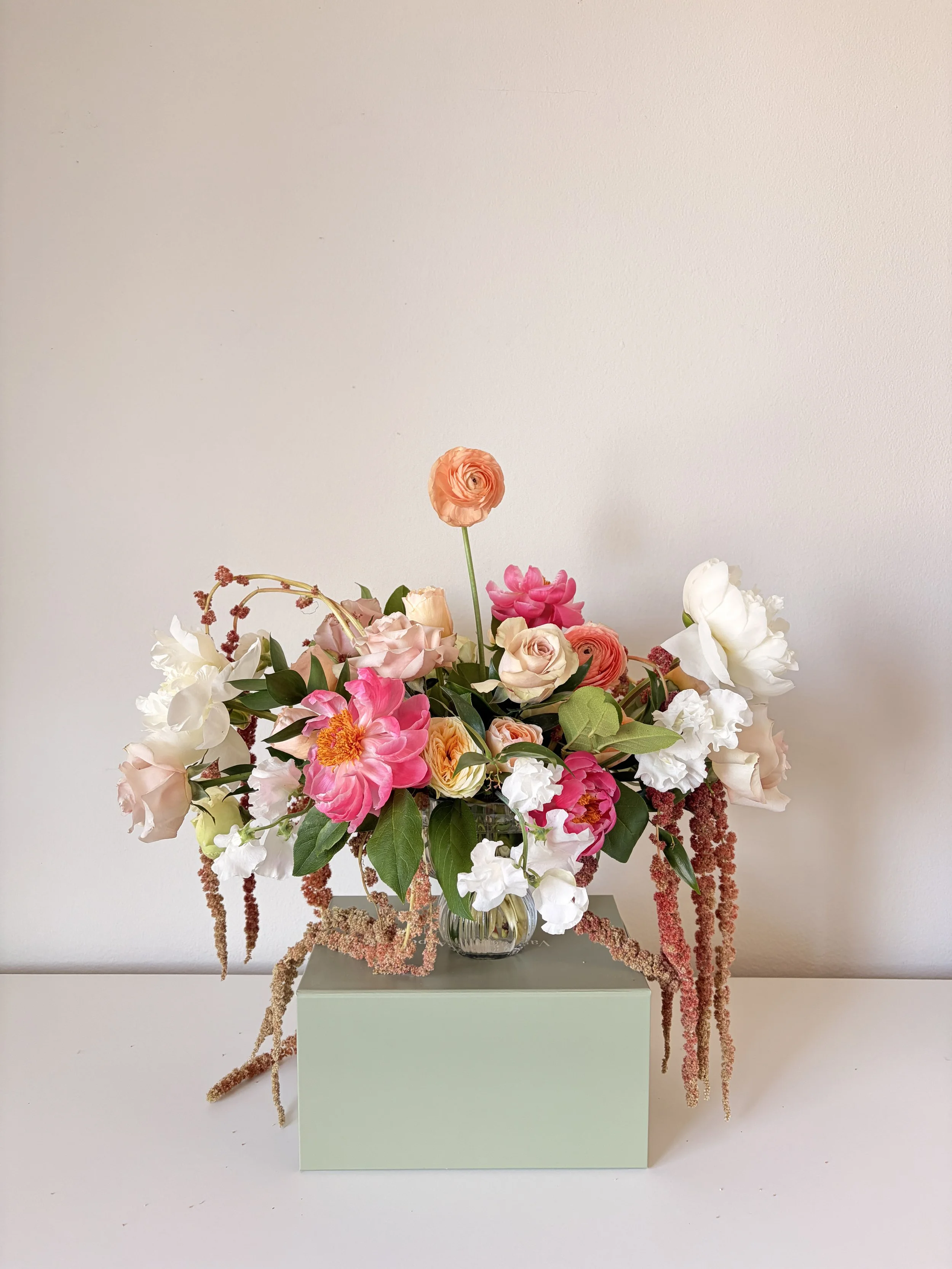 A bouquet of various pink, peach, and white flowers in a clear glass vase on a square pastel green pedestal against a plain light-colored wall.
