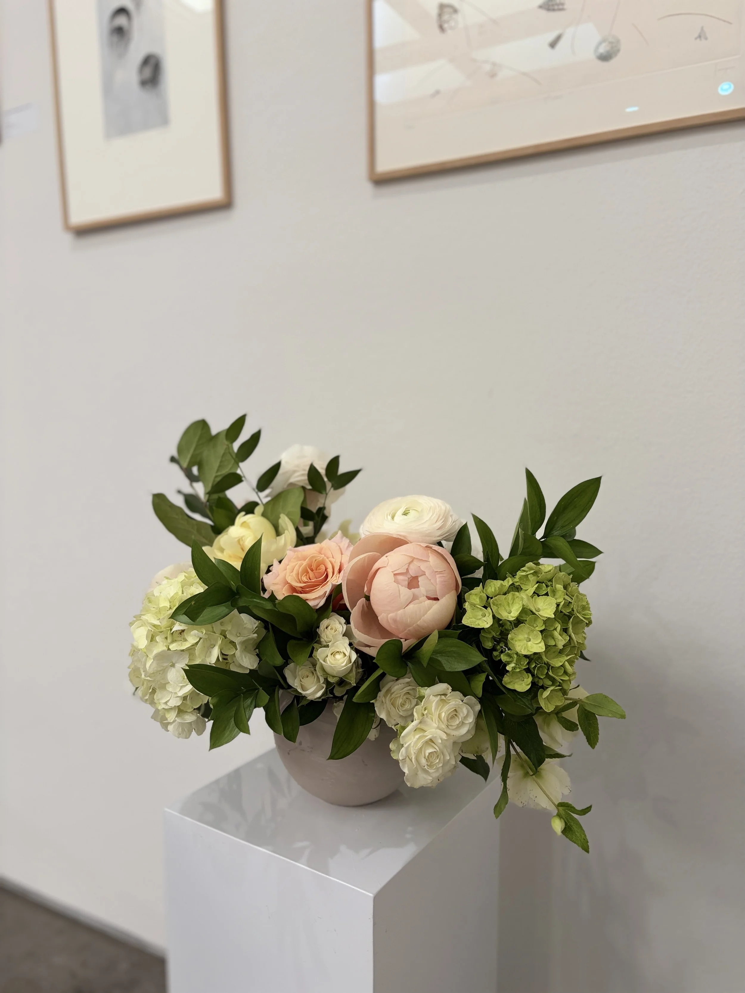 A floral arrangement with white roses, pink peonies, green hydrangeas, and assorted greenery in a white vase on display against a white wall with framed artwork.