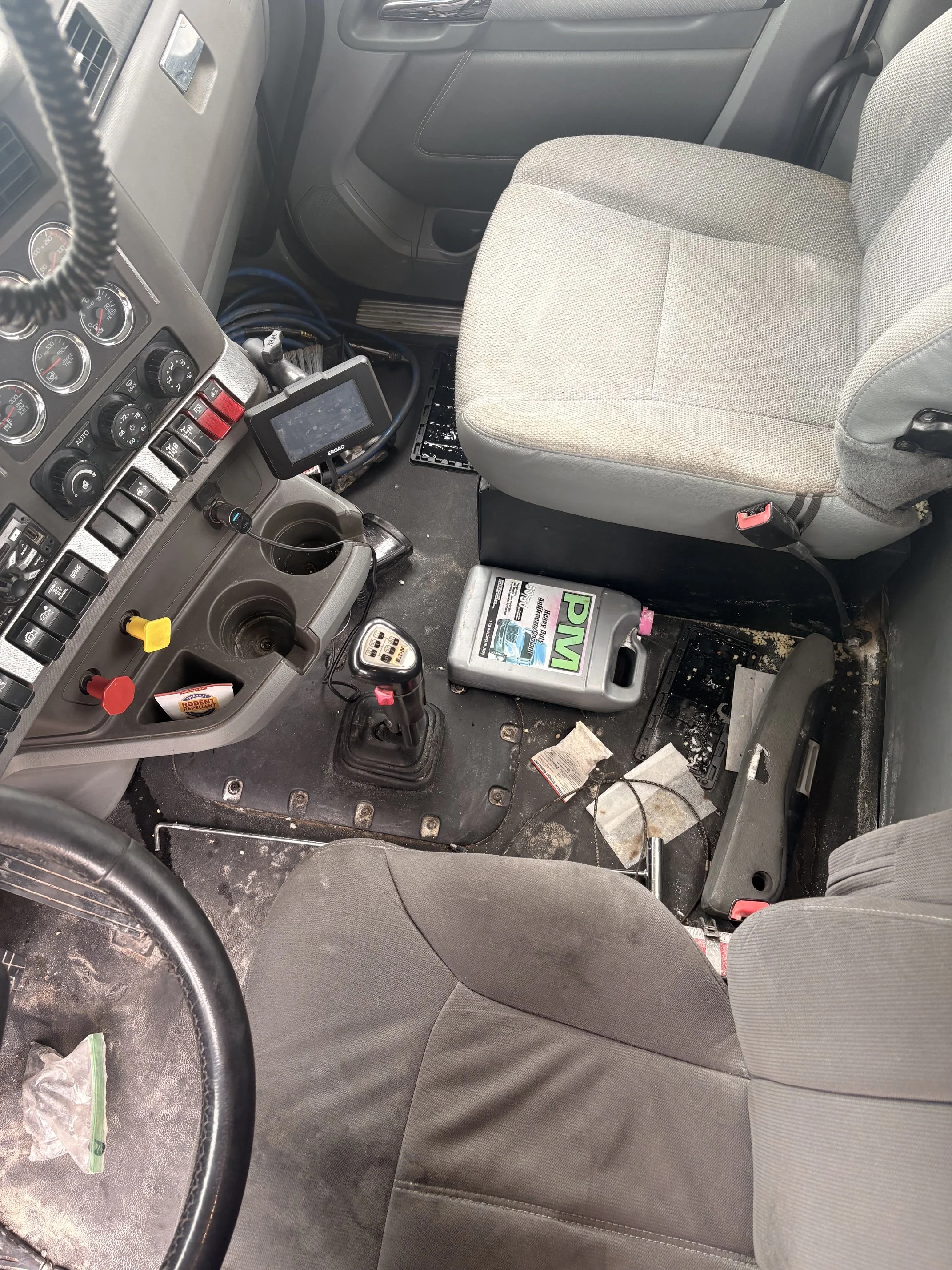 Interior of a vehicle cockpit with the driver's seat, dashboard, several gauges, steering wheel, and various control buttons. The floor is cluttered with debris, paper, and miscellaneous items, and the passenger seat is visible on the right.