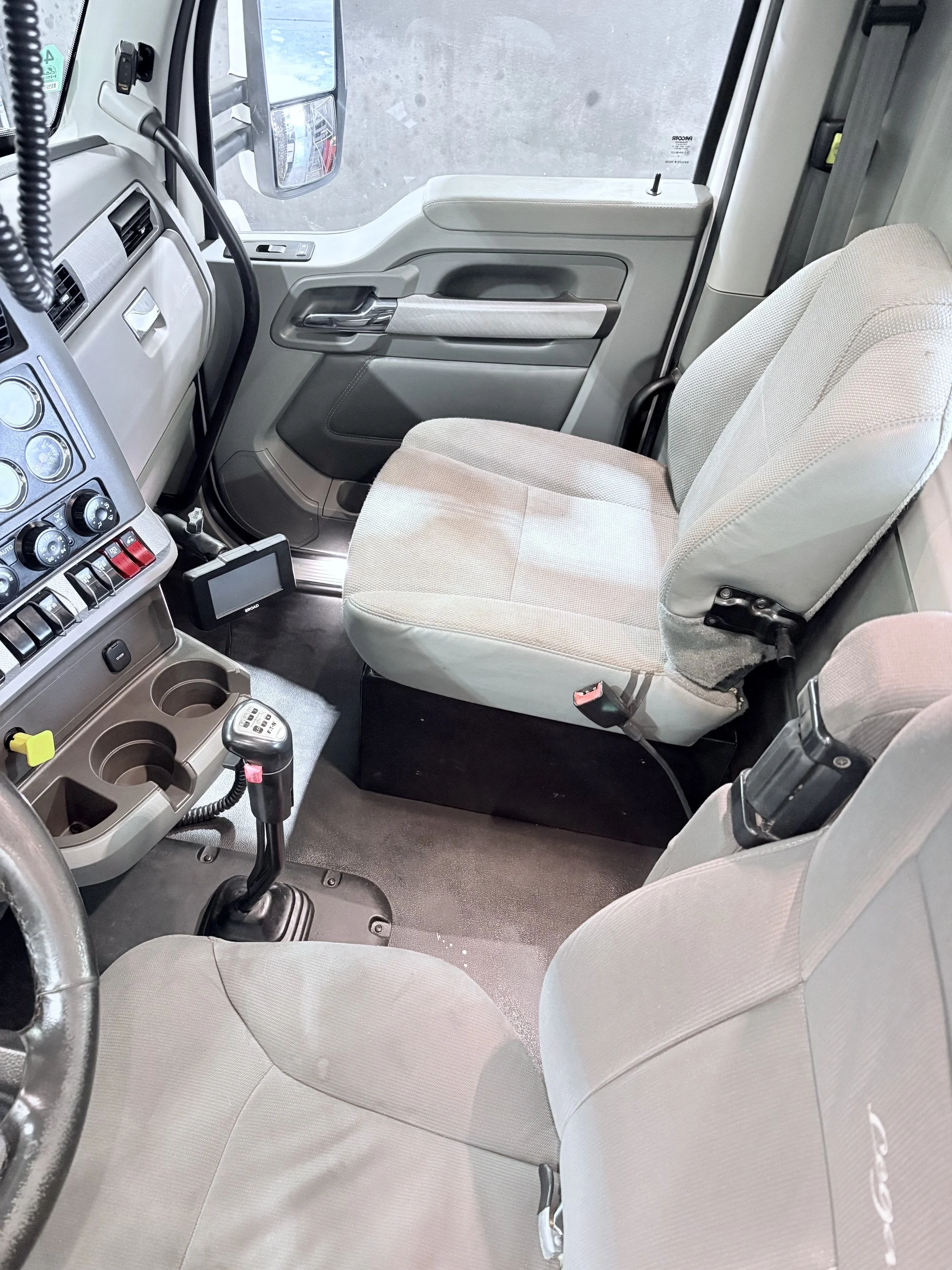 Inside the cabin of a commercial truck, showing the driver’s seat, steering wheel, gear shift, dashboard, and passenger seat, with some control buttons and a small electronic device near the gear shift.