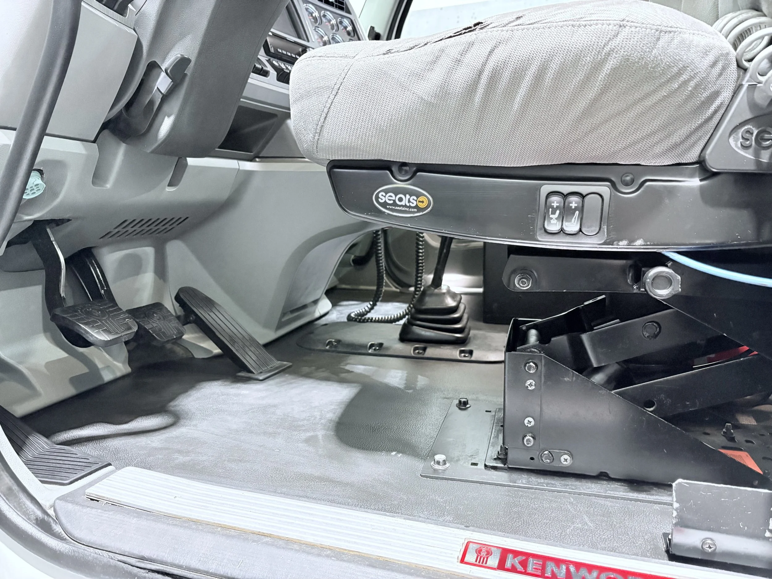 Interior of a bus driver’s seat showing the seat with a gray fabric cover, control buttons on the side, and driver’s pedals including accelerator and brake.