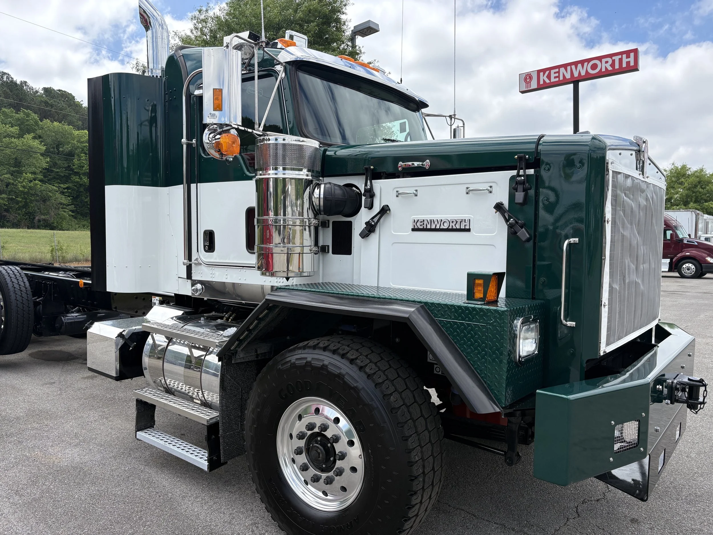 A green and white semi-truck parked outdoors with a Kenworth sign in the background, under a partly cloudy sky.