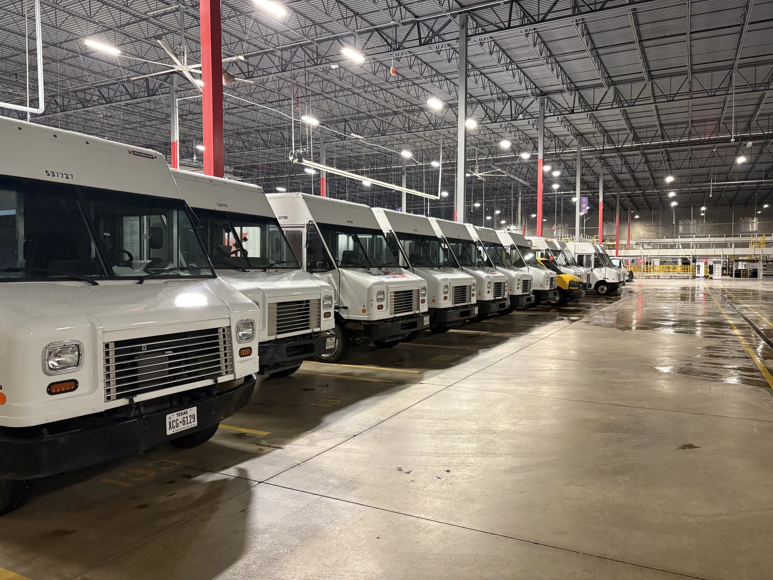 A large indoor space with a row of white delivery trucks parked inside a warehouse or distribution center, with a high ceiling and bright overhead lighting.