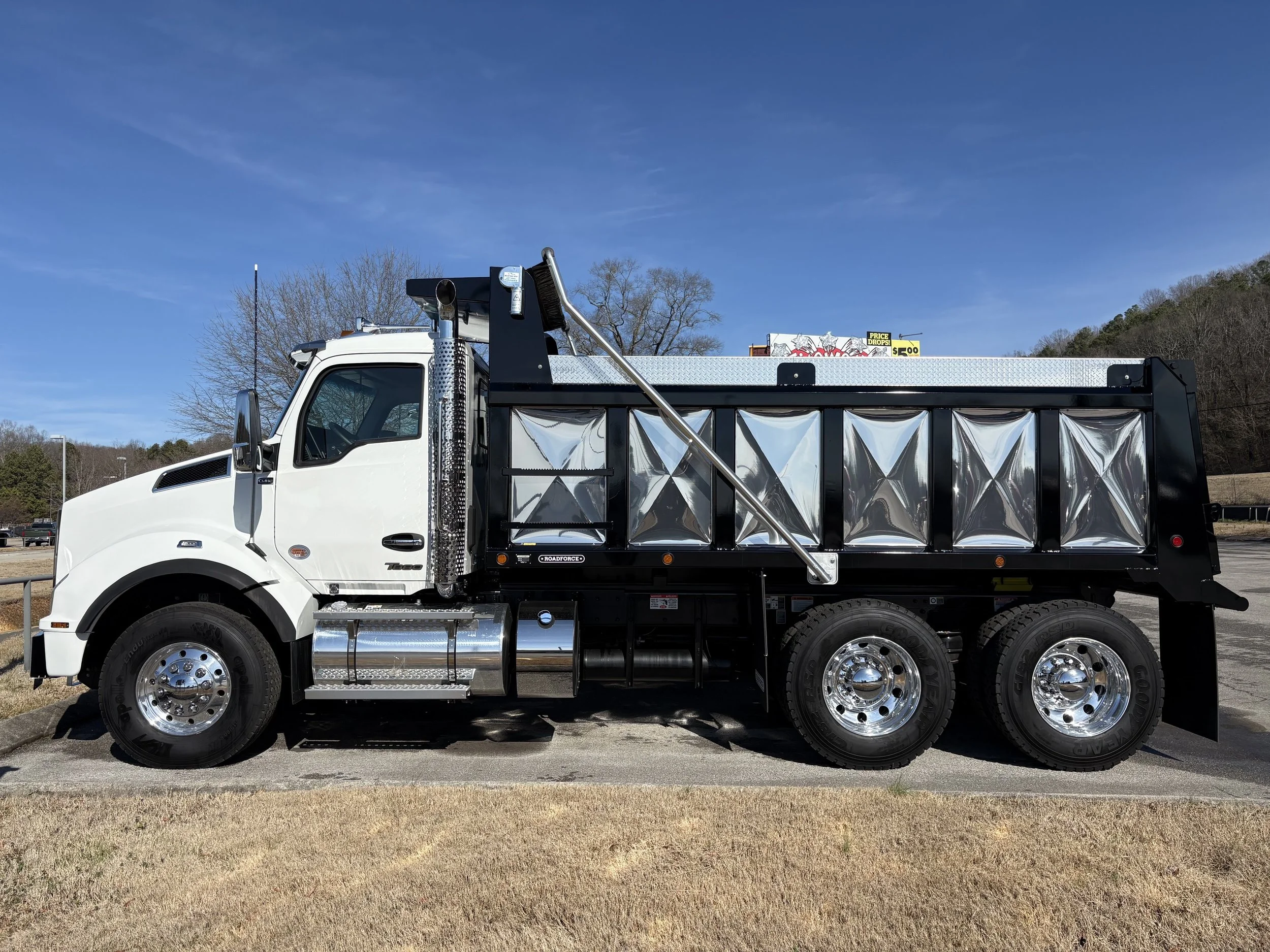 A white and black flatbed truck parked on a parking lot with a blue sky and trees in the background.