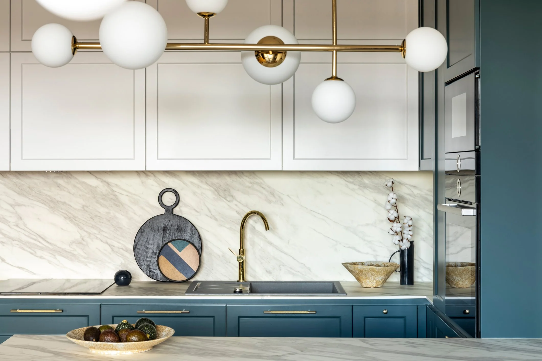 Modern kitchen with blue cabinetry, gold fixtures, white marble backsplash, and decorative objects including a bowl of avocados, black ball, and black vase with white cotton stems.