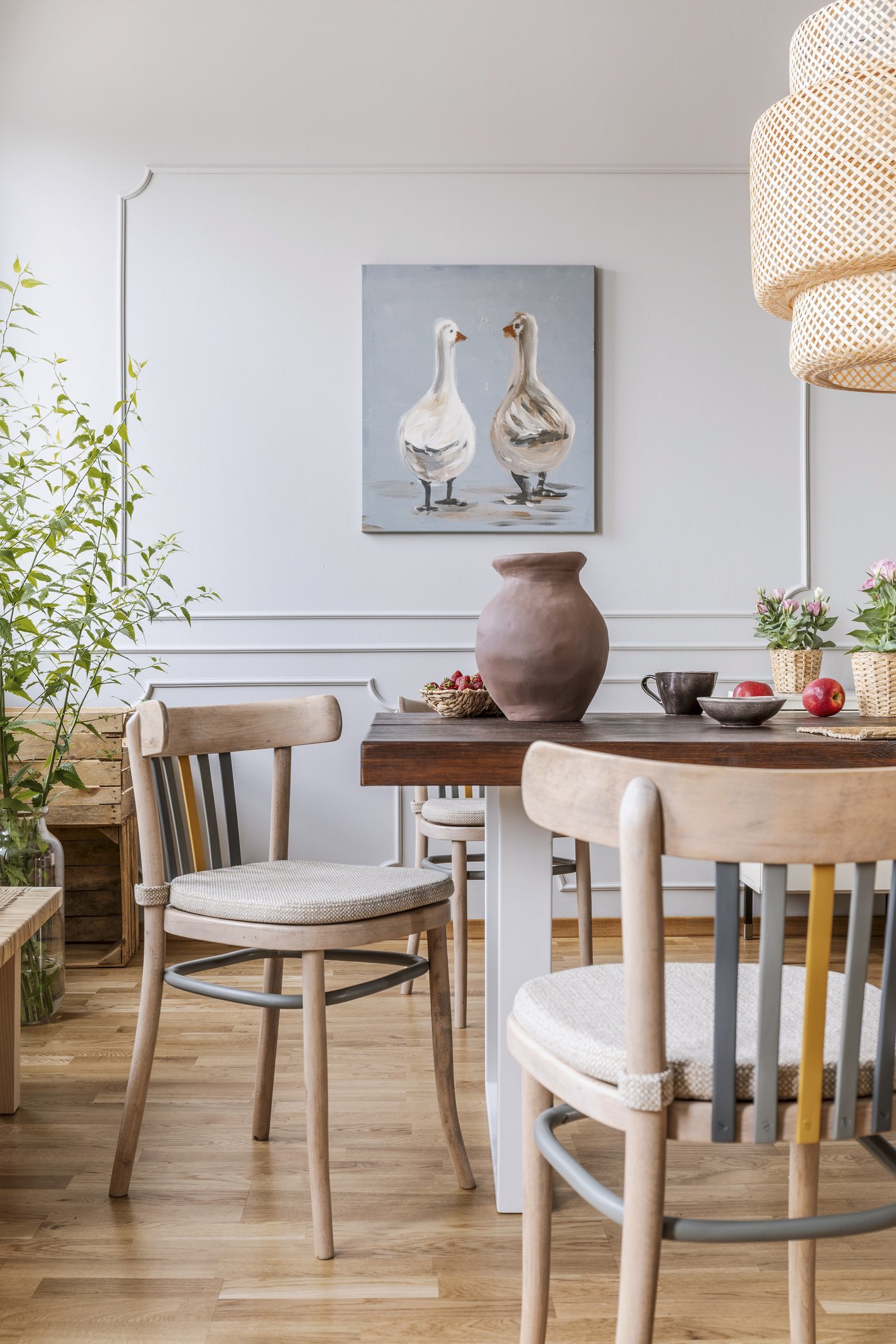 A cozy dining room with a wooden table, light wood chairs, a large brown vase, potted plants, and a painting of two geese on the wall.