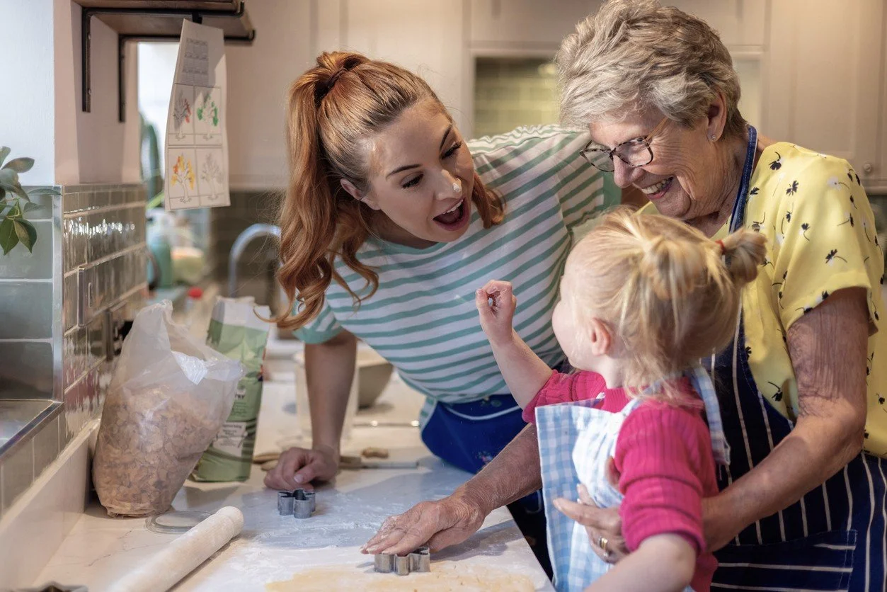 Three females, a young woman, an elderly woman, and a young girl, baking and decorating cookies together in a kitchen. The young woman and elderly woman smile and laugh, while the girl looks at the elderly woman.