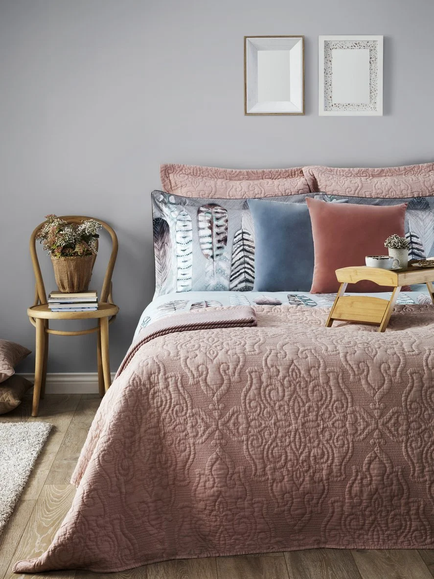A cozy bedroom with a pink quilted bedspread, decorative pillows in pink, blue, and patterned fabrics, and framed mirrors on a light gray wall. A small wooden tray holds a flower-vase and a cup. There is a wooden chair with a potted plant and books beside the bed.